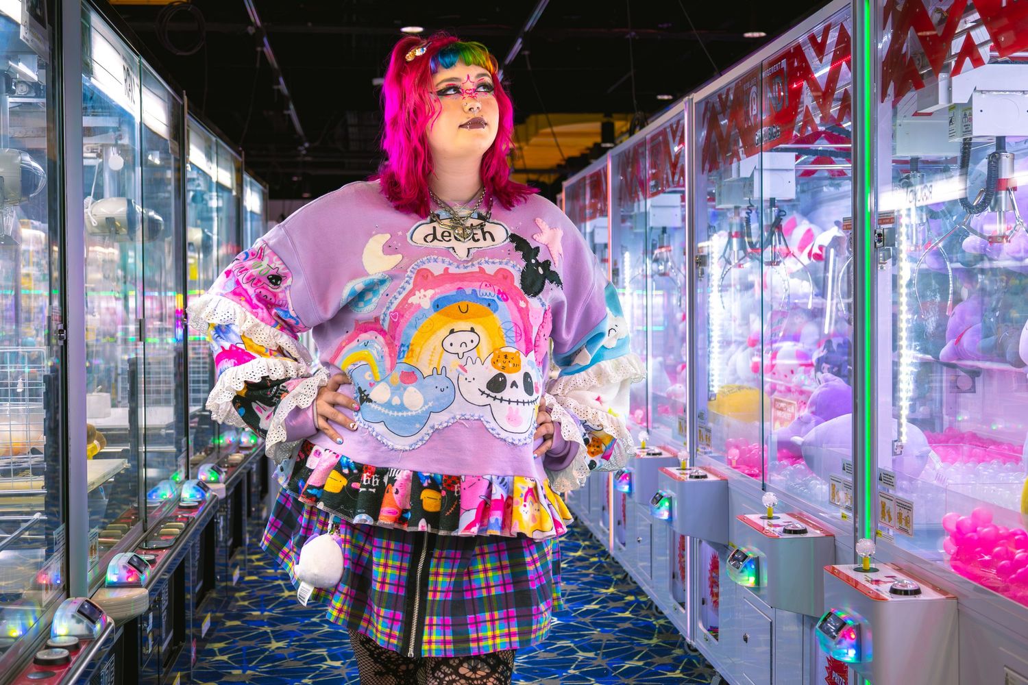 Woman with pink hair in colorful kawaii sweater poses in arcade claw machine hall wearing plaid skirt.