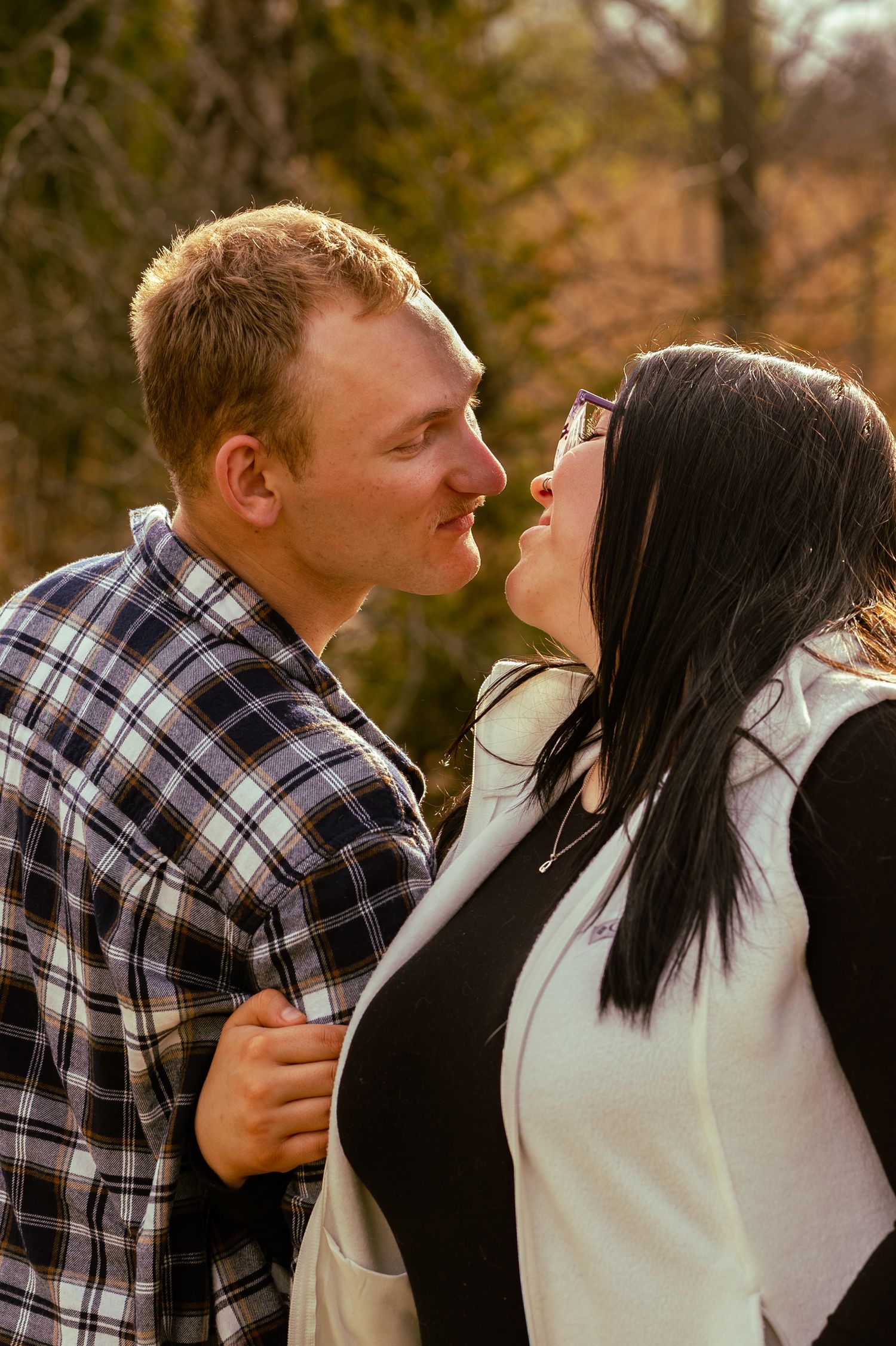 A couple shares a romantic moment outdoors, with the man leaning in for a kiss with a woman wearing glasses.