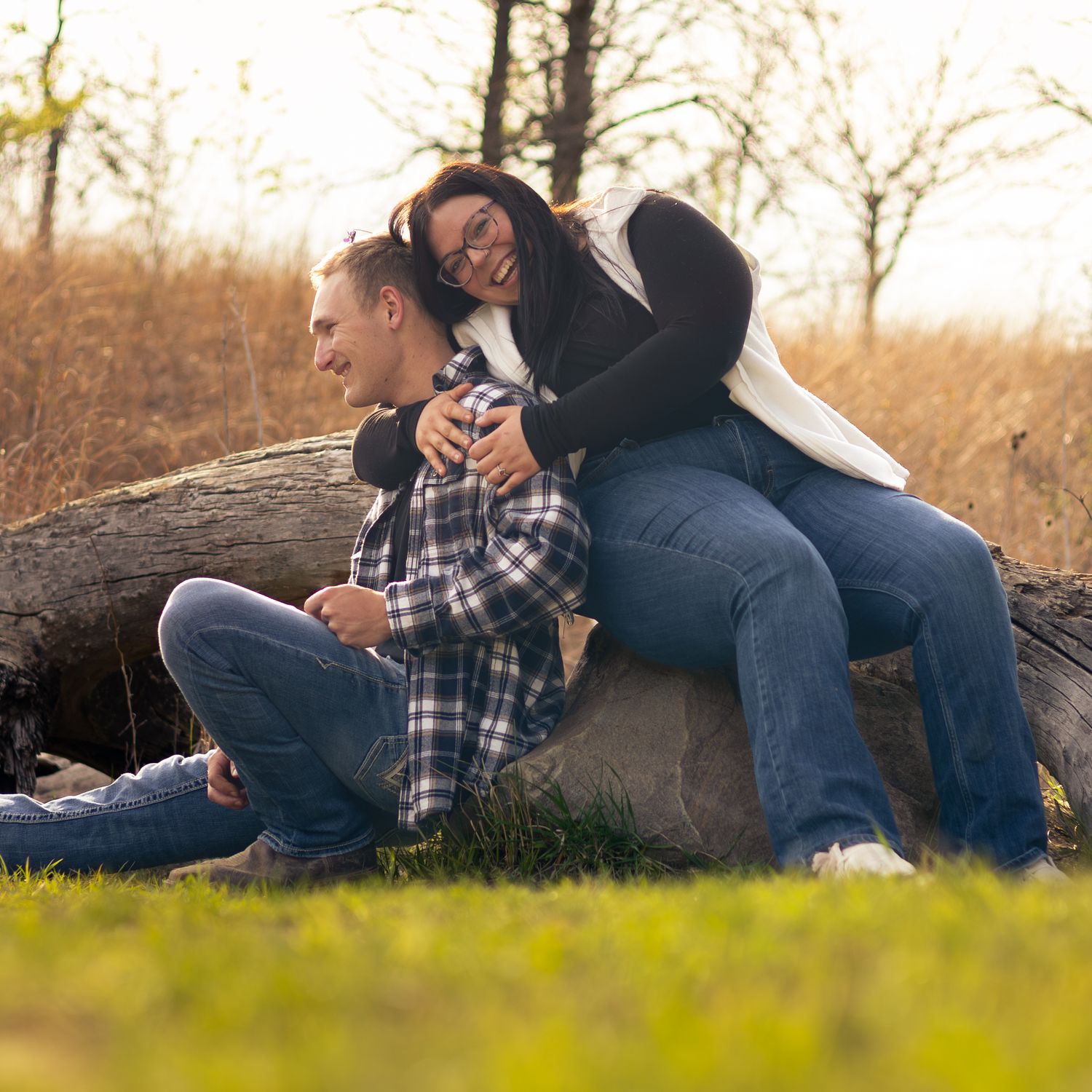 A couple sits on a fallen log outdoors, smiling and embracing in a casual autumn engagement photo session.
