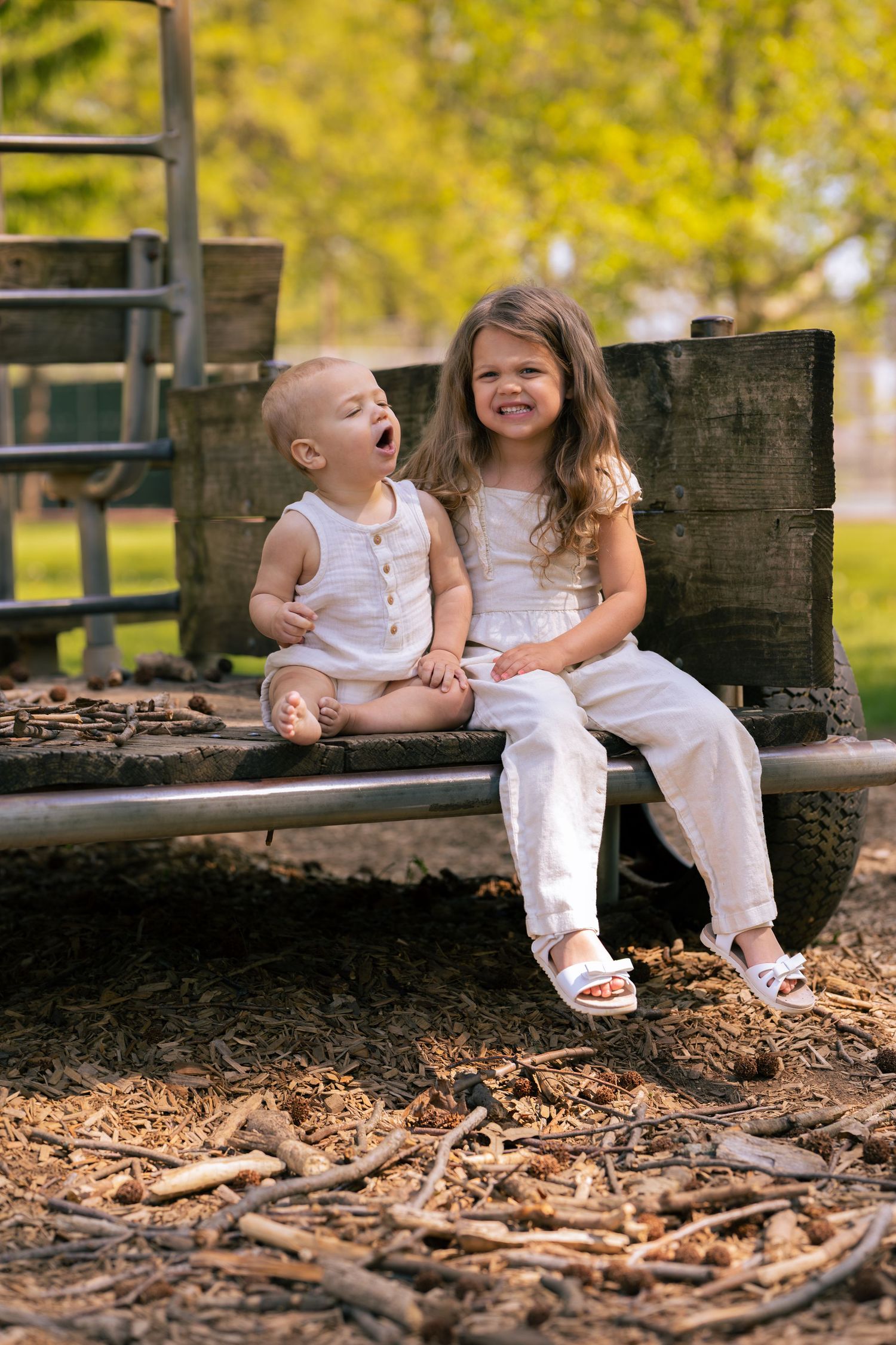 Two young children in white outfits laughing and sitting together on a rustic wooden bench outdoors.