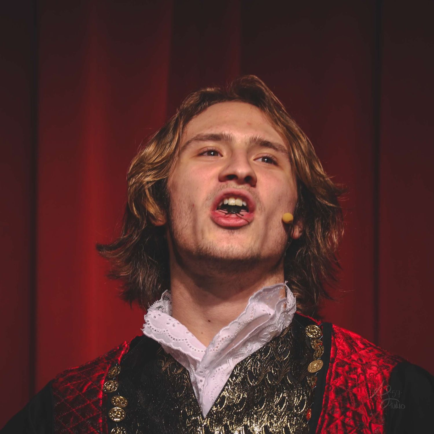 Performer in ornate red costume and theatrical makeup against red curtain backdrop.