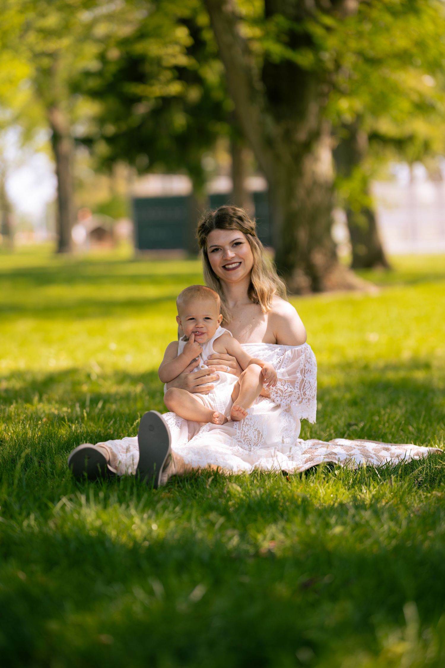 Young mother in white lace dress sitting on grass in park holding smiling baby on sunny day.
