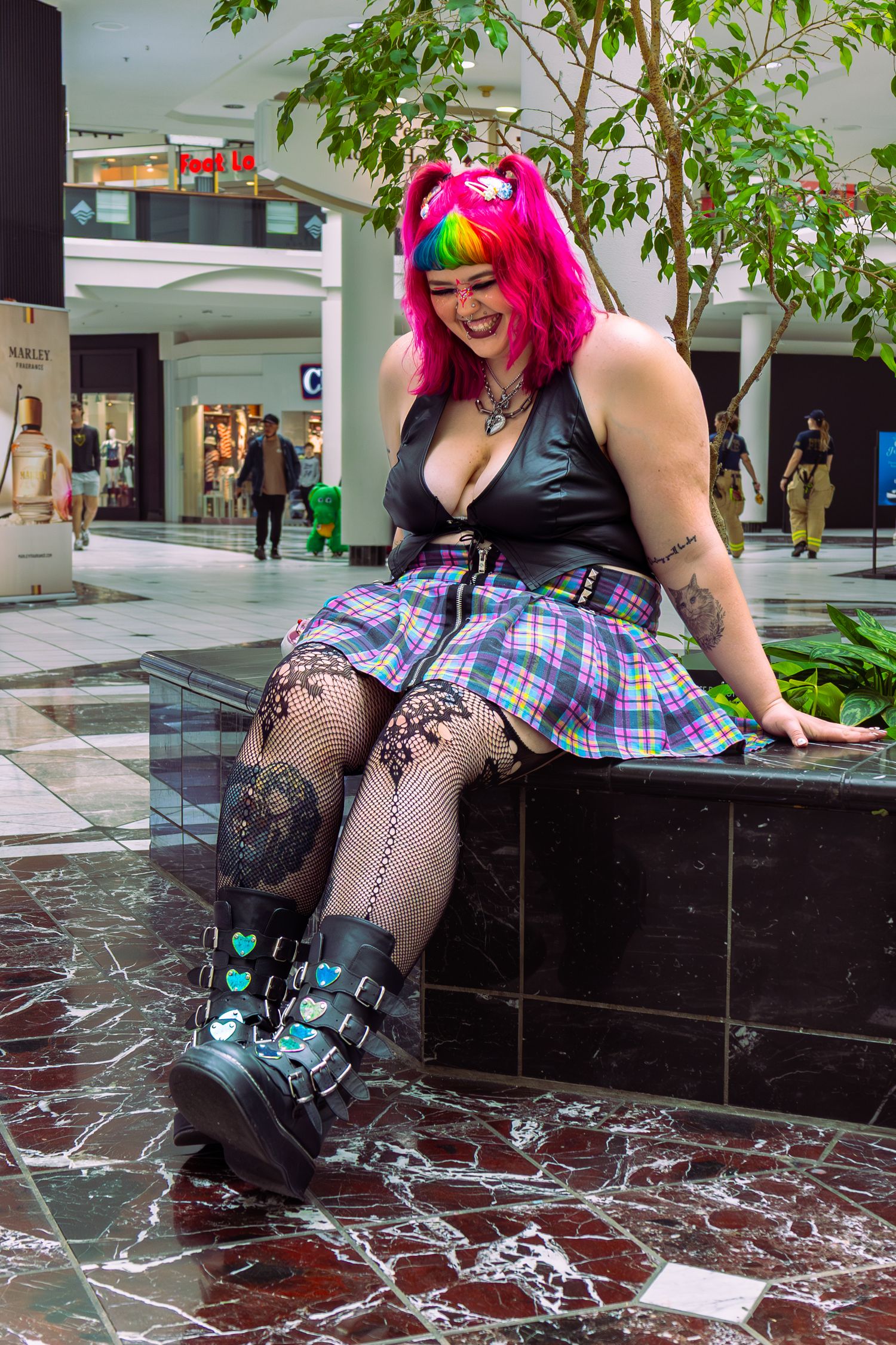 Woman in rainbow wig, plaid skirt, fishnet tights, and platform boots sitting in a shopping mall.