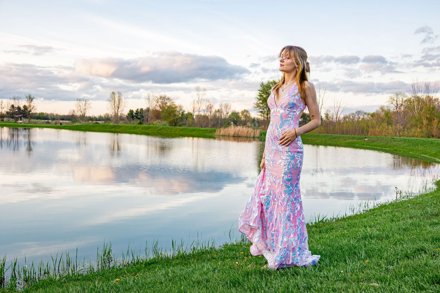 Person in floral dress standing by lake during sunset with cloudy sky.