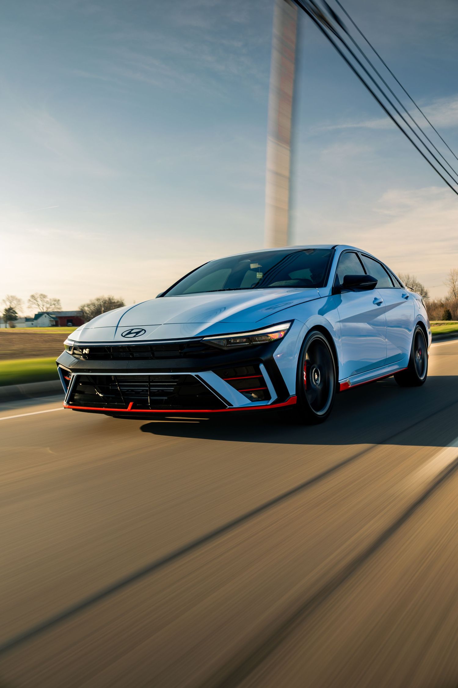 A white Hyundai Elantra N sedan with red accents speeds down a road at sunset in a dynamic motion-blur shot.