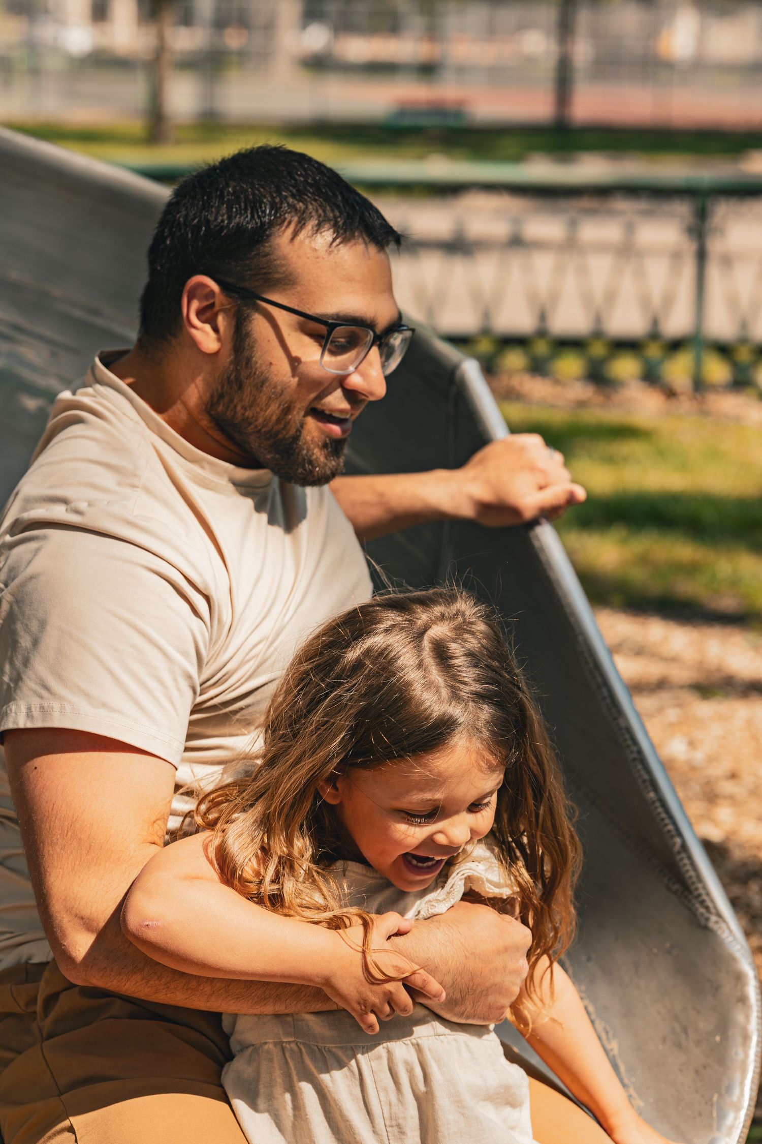 Father and daughter laughing together on a playground slide on a sunny day.