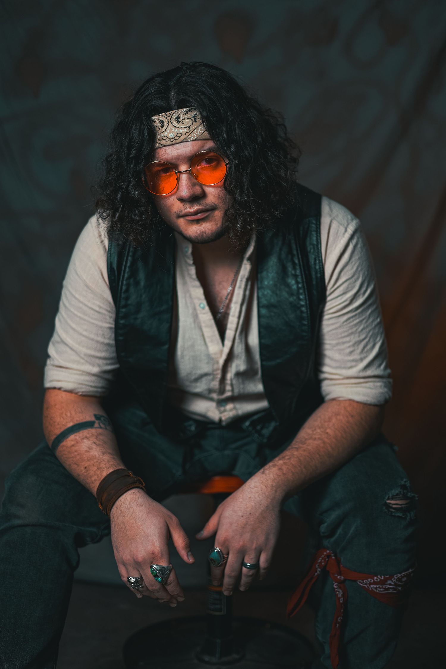 Portrait of person in hippie attire sitting casually against dark background.