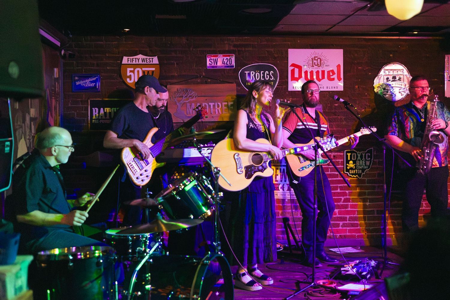 Live band performing on stage in a dimly lit bar, featuring guitar, drums, and saxophone players.
