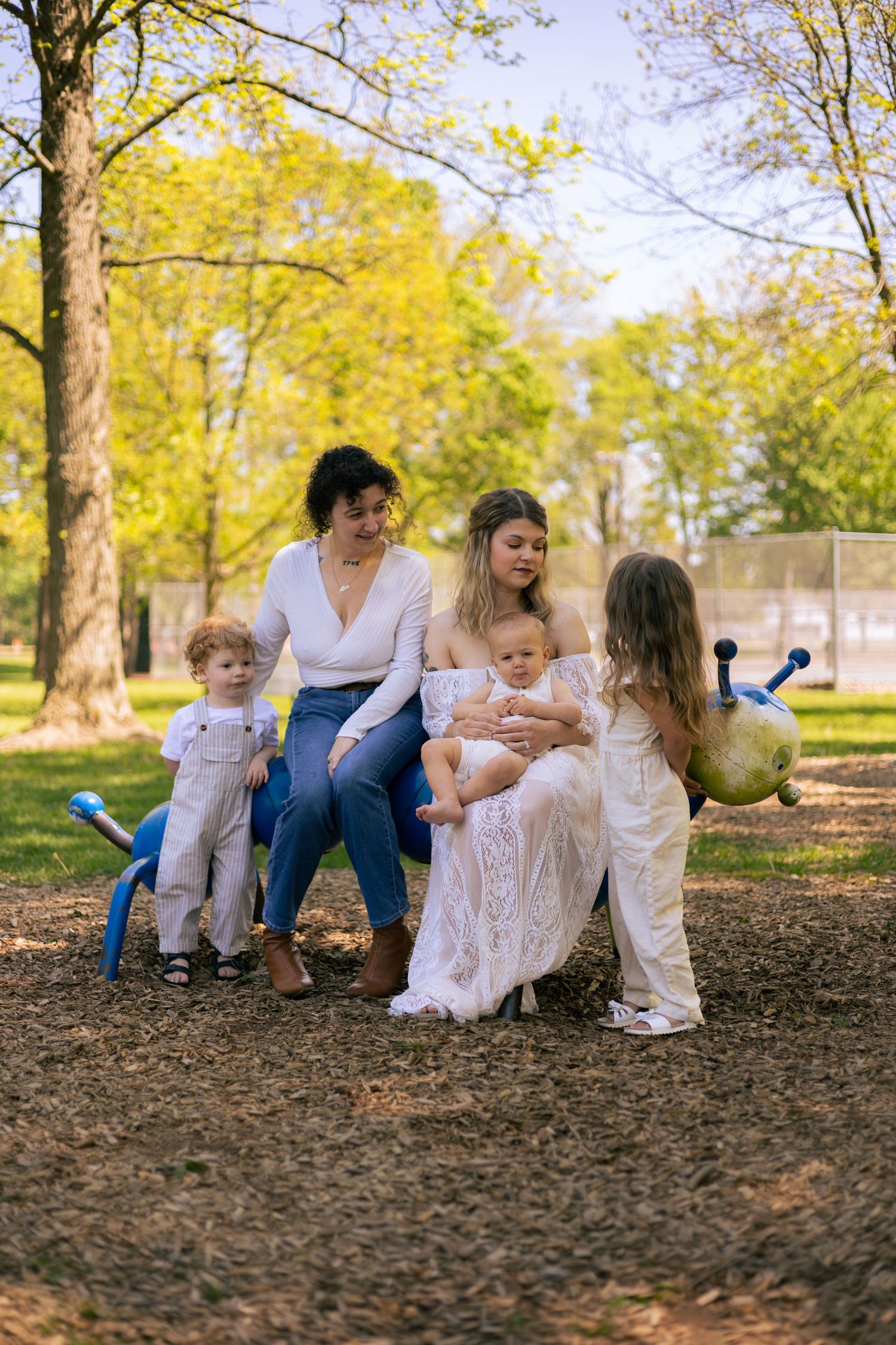 Two women sitting in a park with three young children, surrounded by trees with spring foliage.