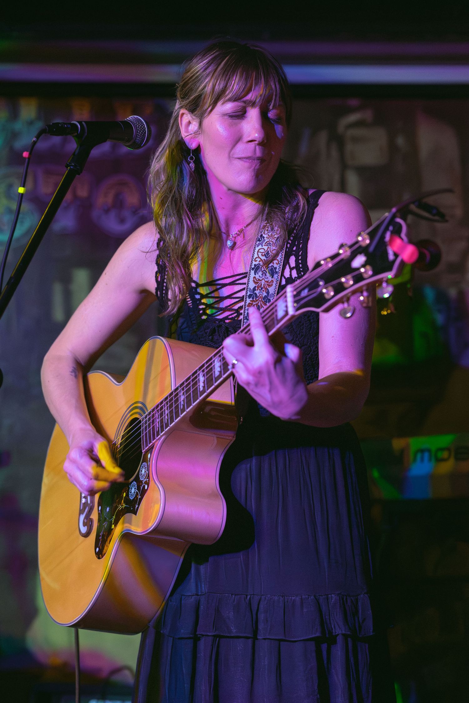 Woman playing acoustic guitar on stage at a live music venue, performing under colorful stage lighting.