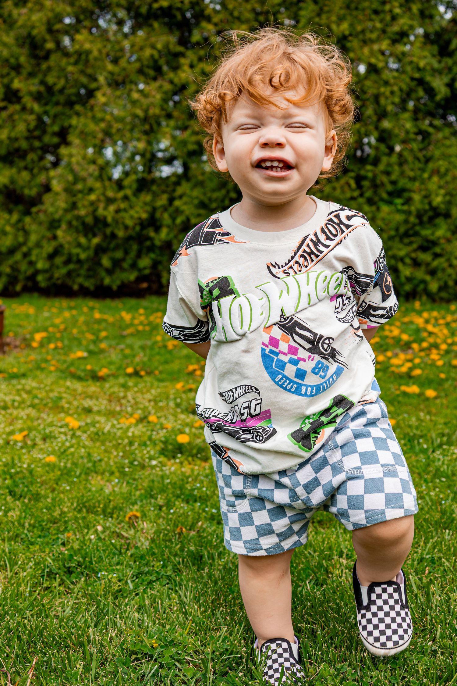 Child in checkered shorts and graphic tee plays on green grass with yellow dandelions in background.