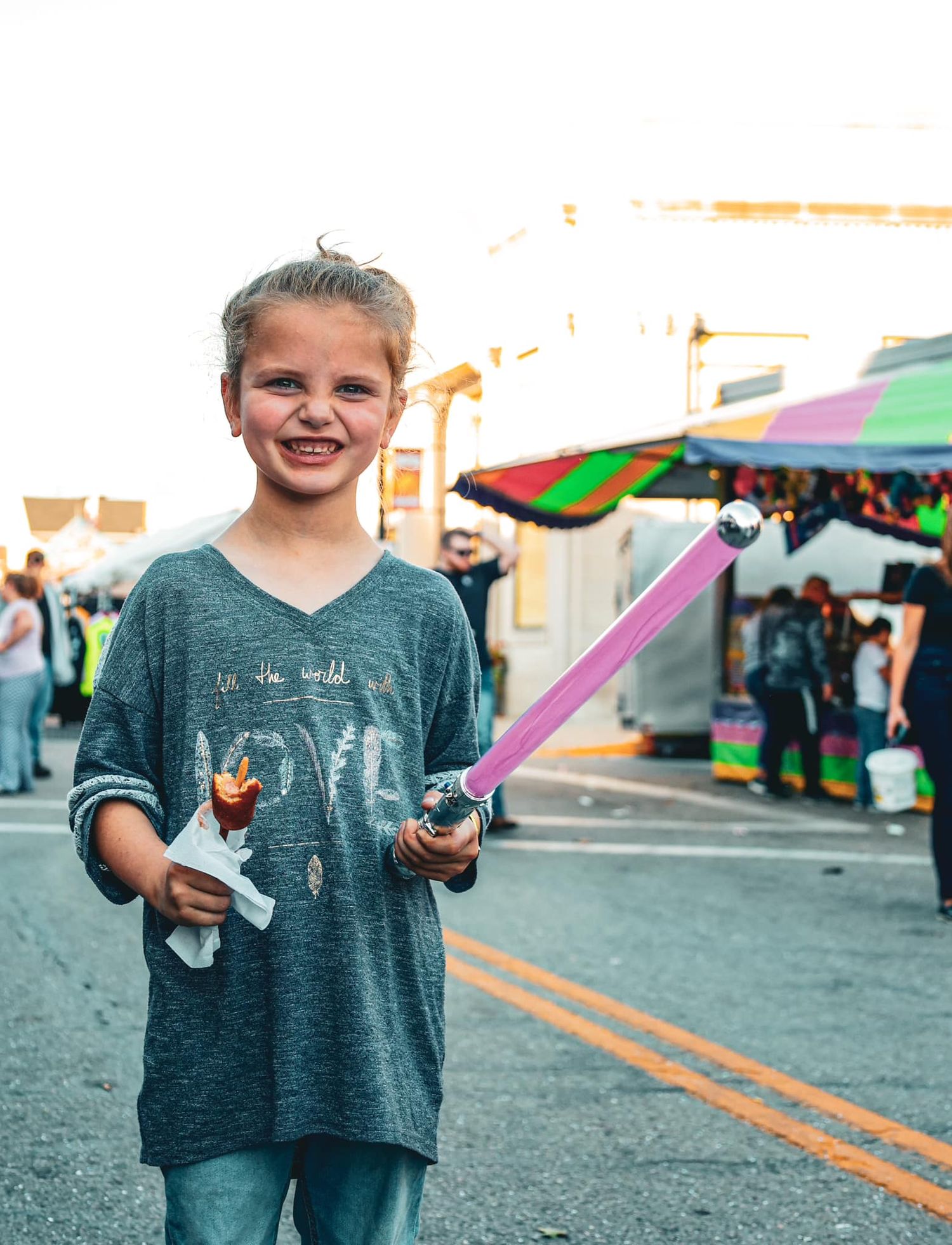 Child holding a pink toy at a carnival with colorful tents in the background.
