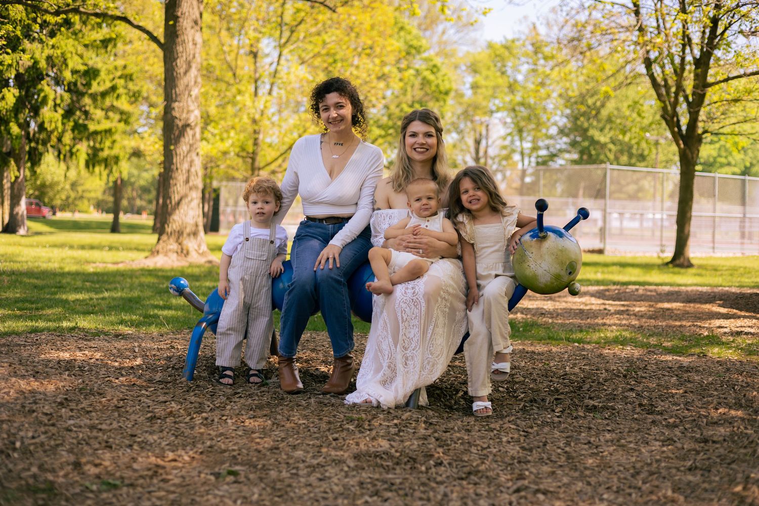 Two women sit with three young children in a sunny park, smiling for a family portrait among green trees.