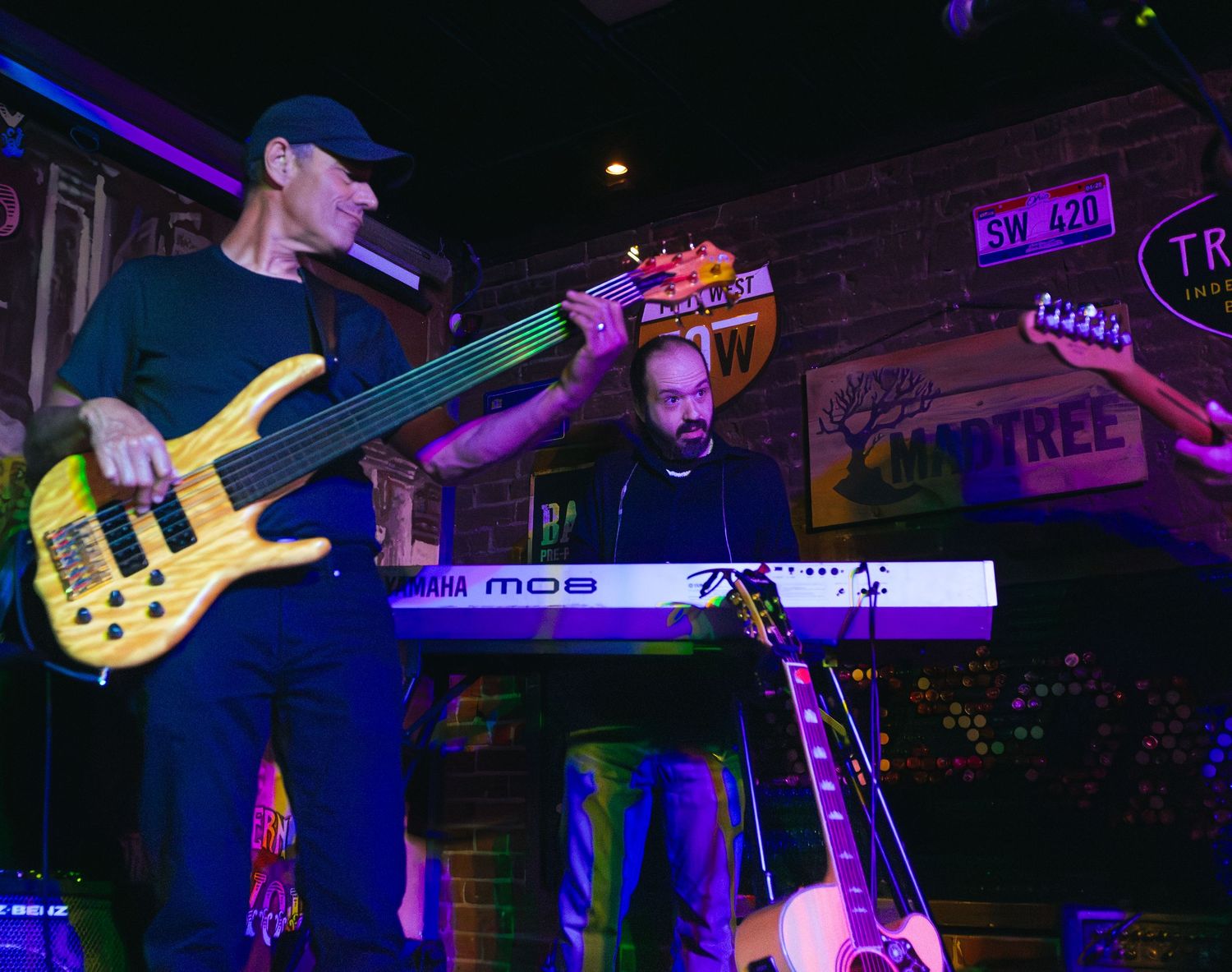 Two musicians perform live, one playing bass guitar and another at a keyboard on a dimly lit bar stage.