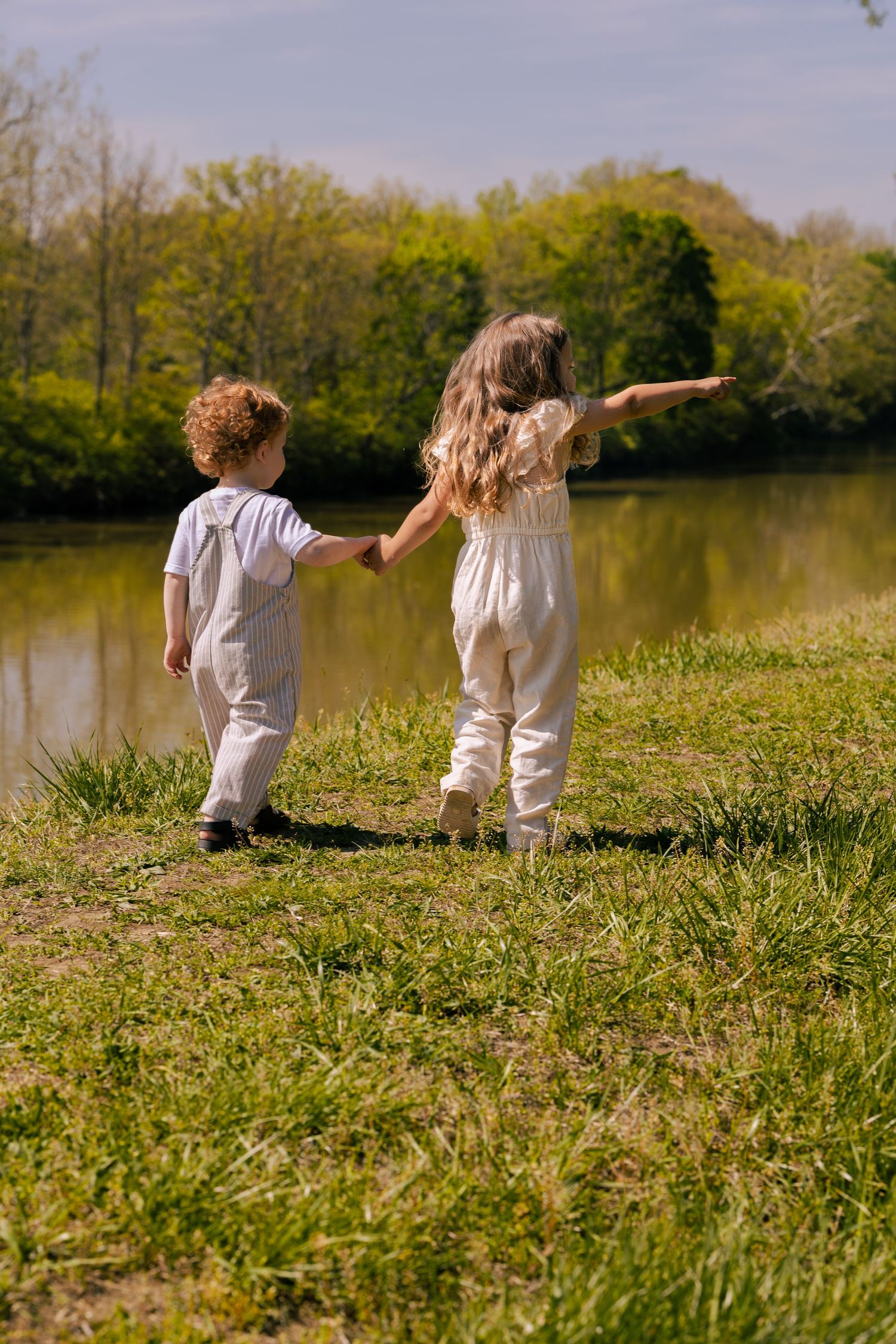 Two young children holding hands near a peaceful pond surrounded by lush green trees on a sunny day.