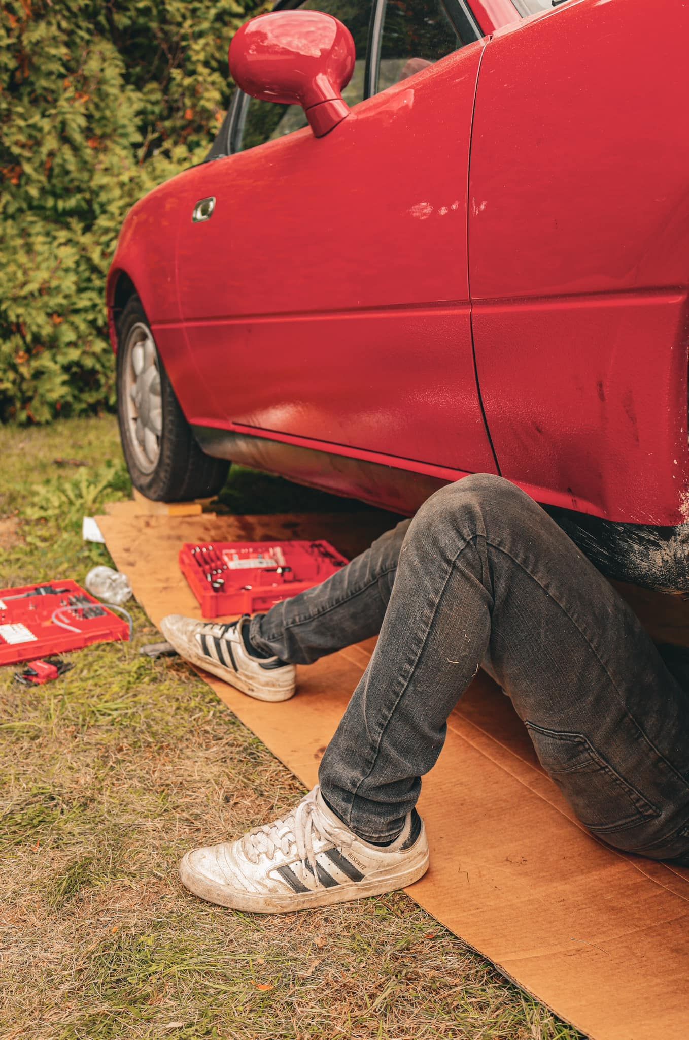Person's legs in jeans and sneakers visible under a red car during maintenance.