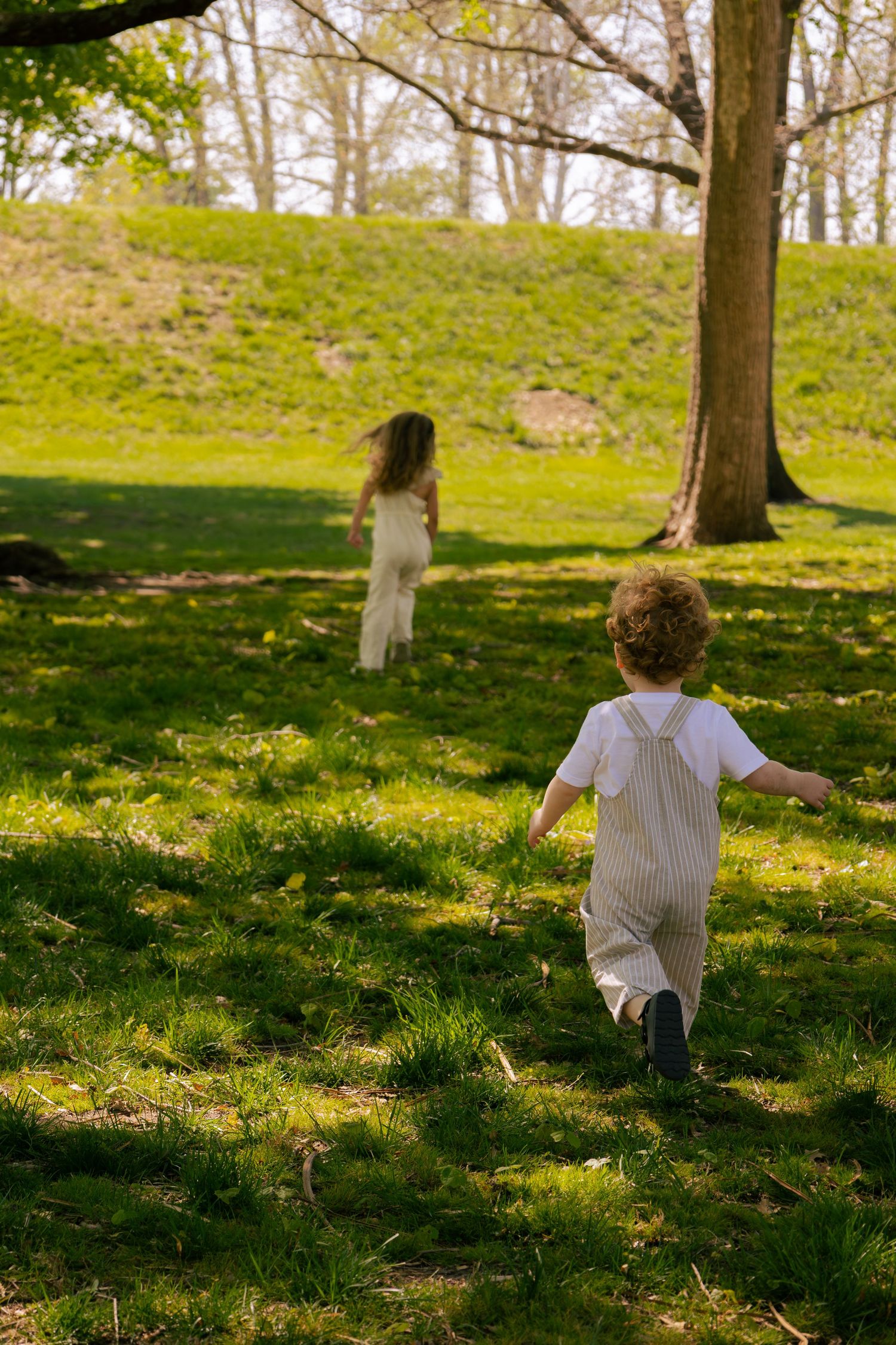 Two children running and playing on lush green grass in a sunny park with trees in the background.