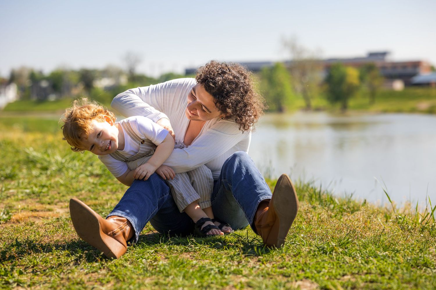 Mother and child playing and laughing together on green grass by a lake on a sunny day.