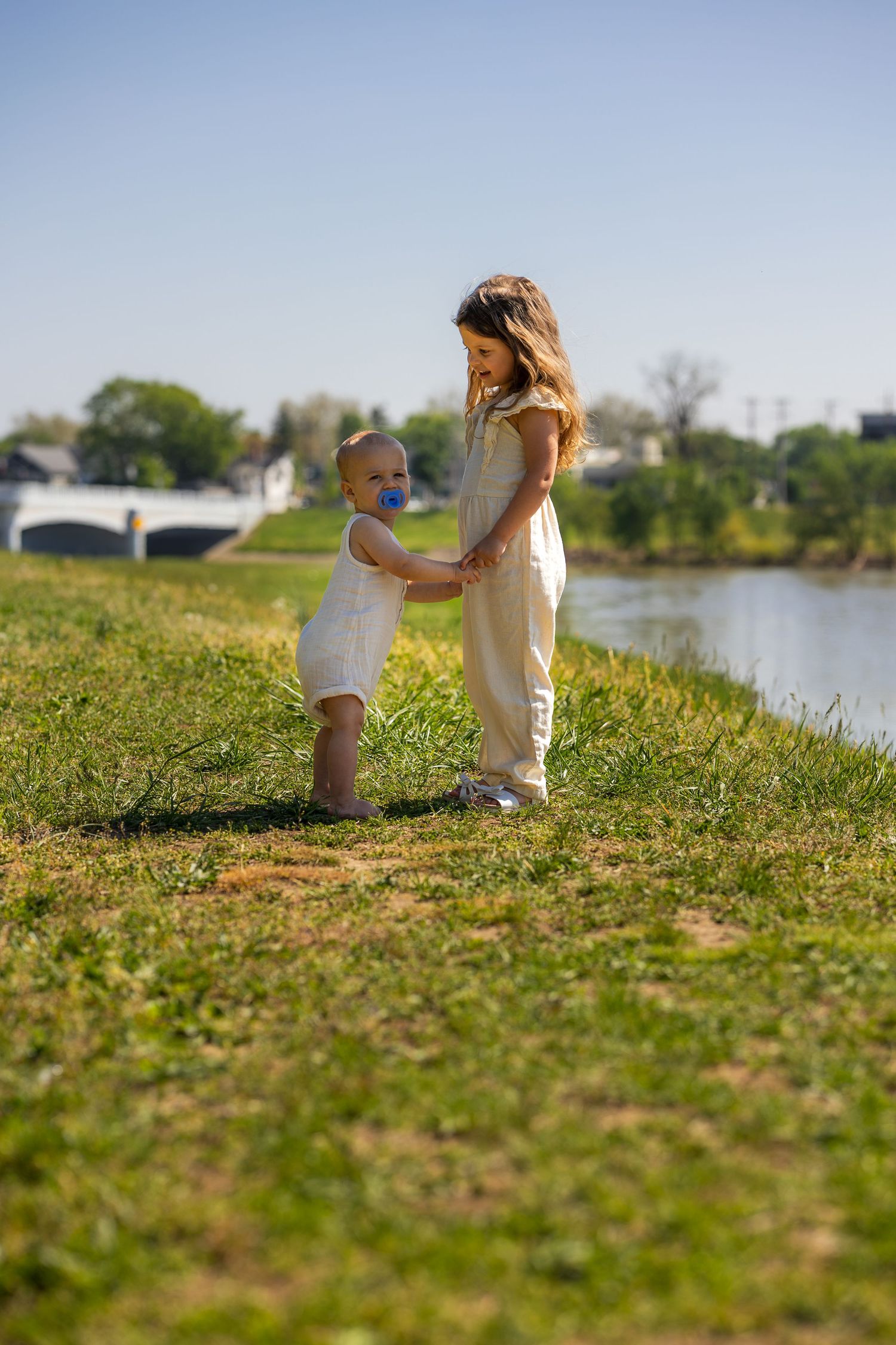 A young girl holds hands with a toddler with a pacifier on a grassy riverbank on a sunny day.