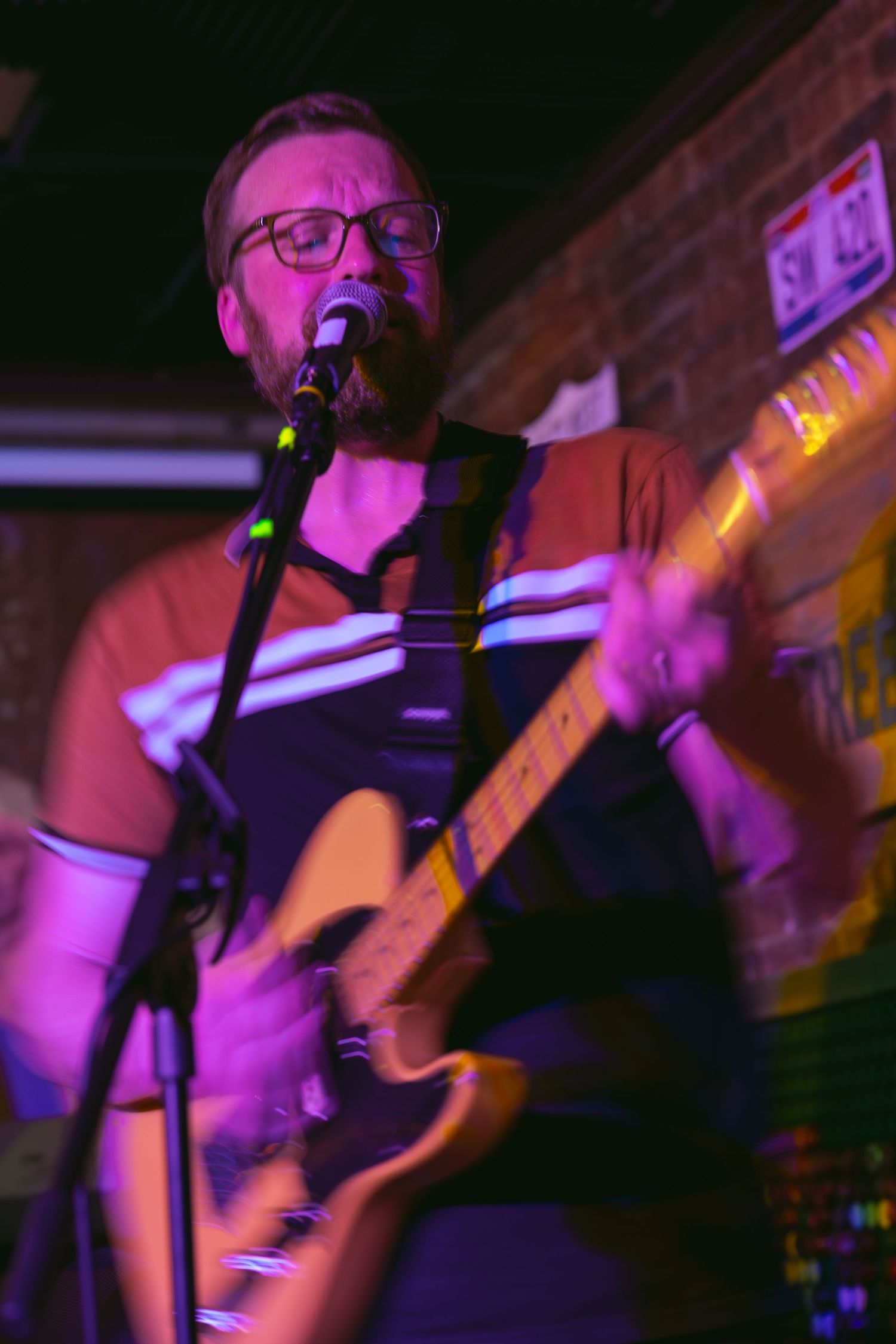 Man with glasses playing electric guitar and singing into microphone on stage under purple lights at a live music venue.