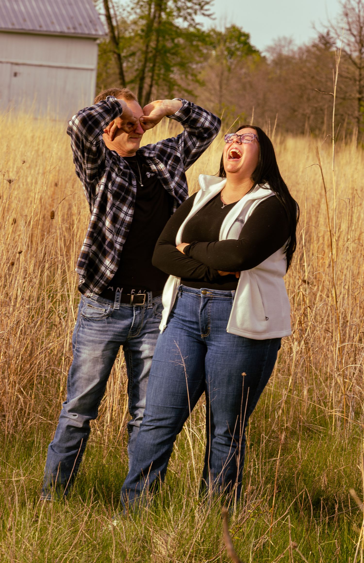 Two people laughing joyfully in a golden field, wearing jeans and casual tops, with a barn and trees in the background.