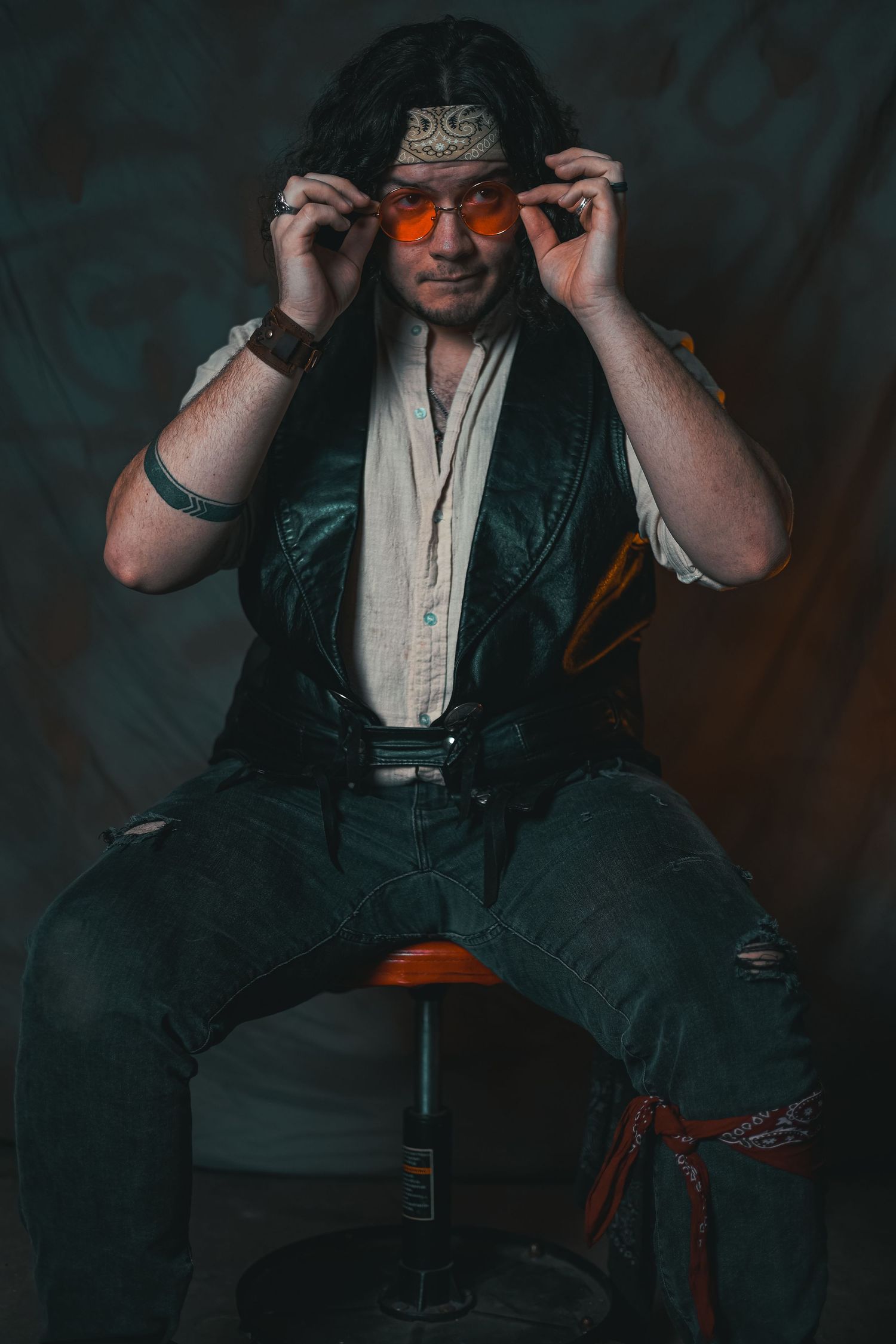 Stylish portrait of someone adjusting sunglasses while sitting on a stool against dark background.
