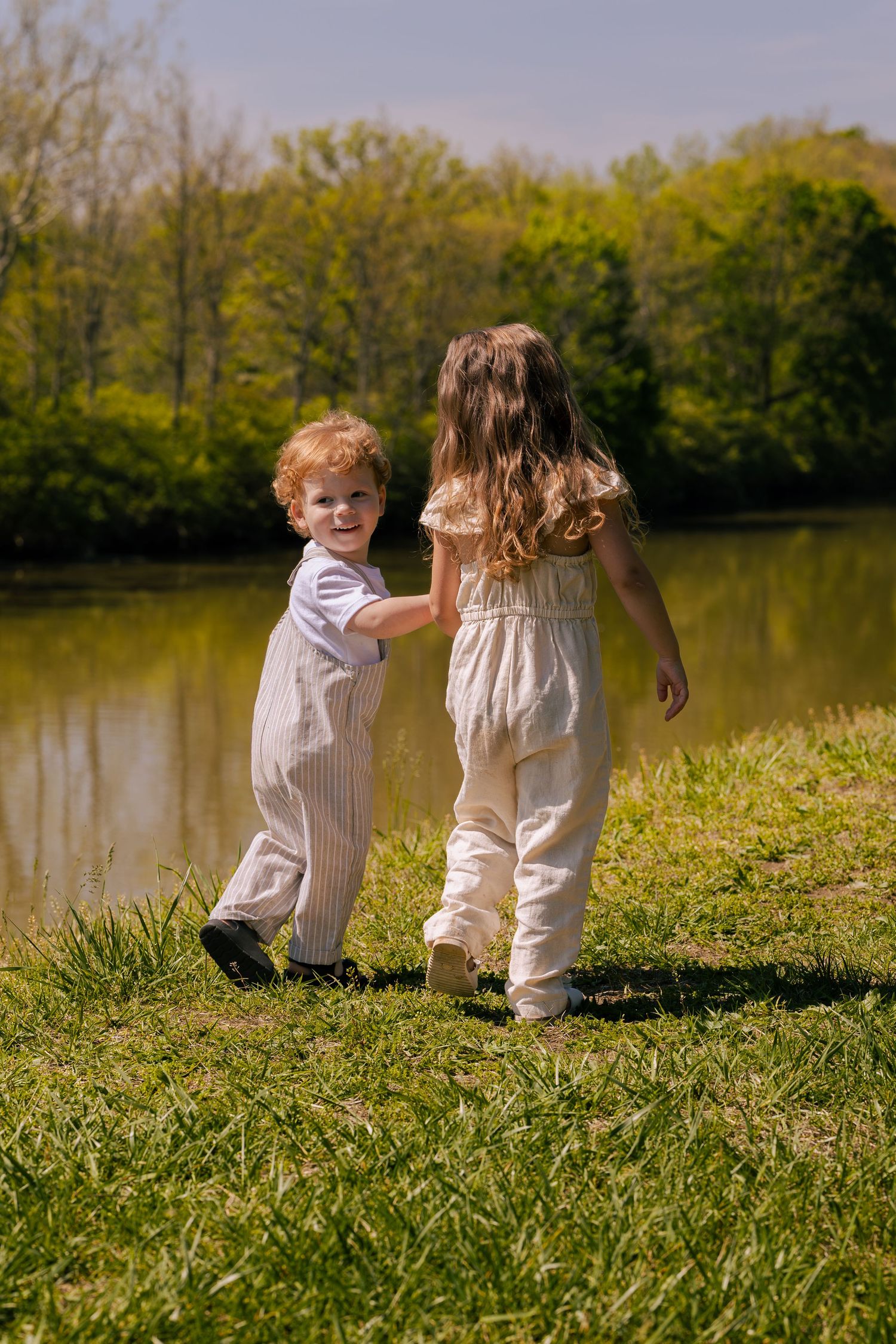 Two young children holding hands near a pond in a sunny park, boy in striped overalls looking back, girl in cream outfit.