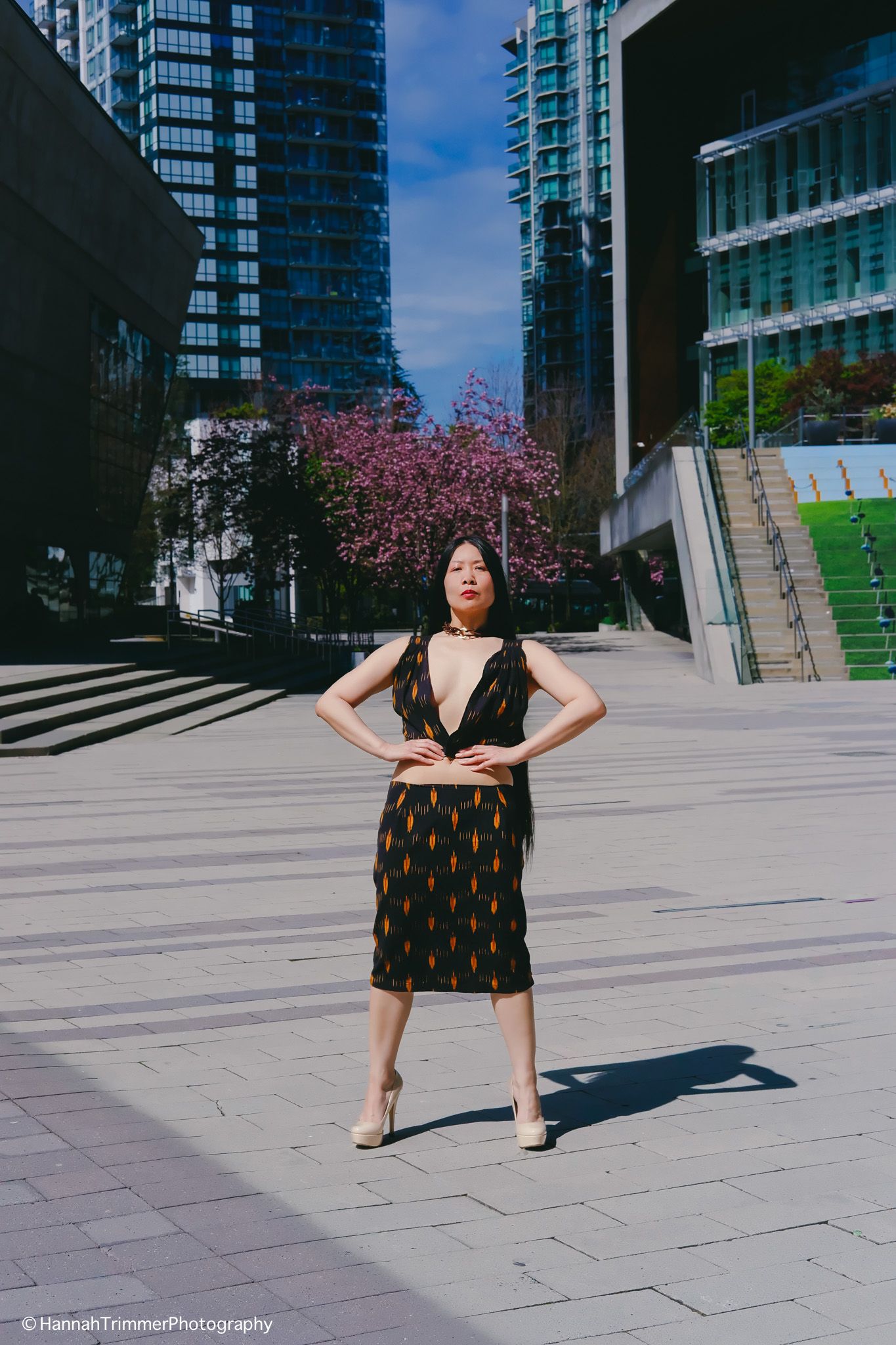 Woman in black patterned dress with hands on hips poses confidently in sunny urban plaza.