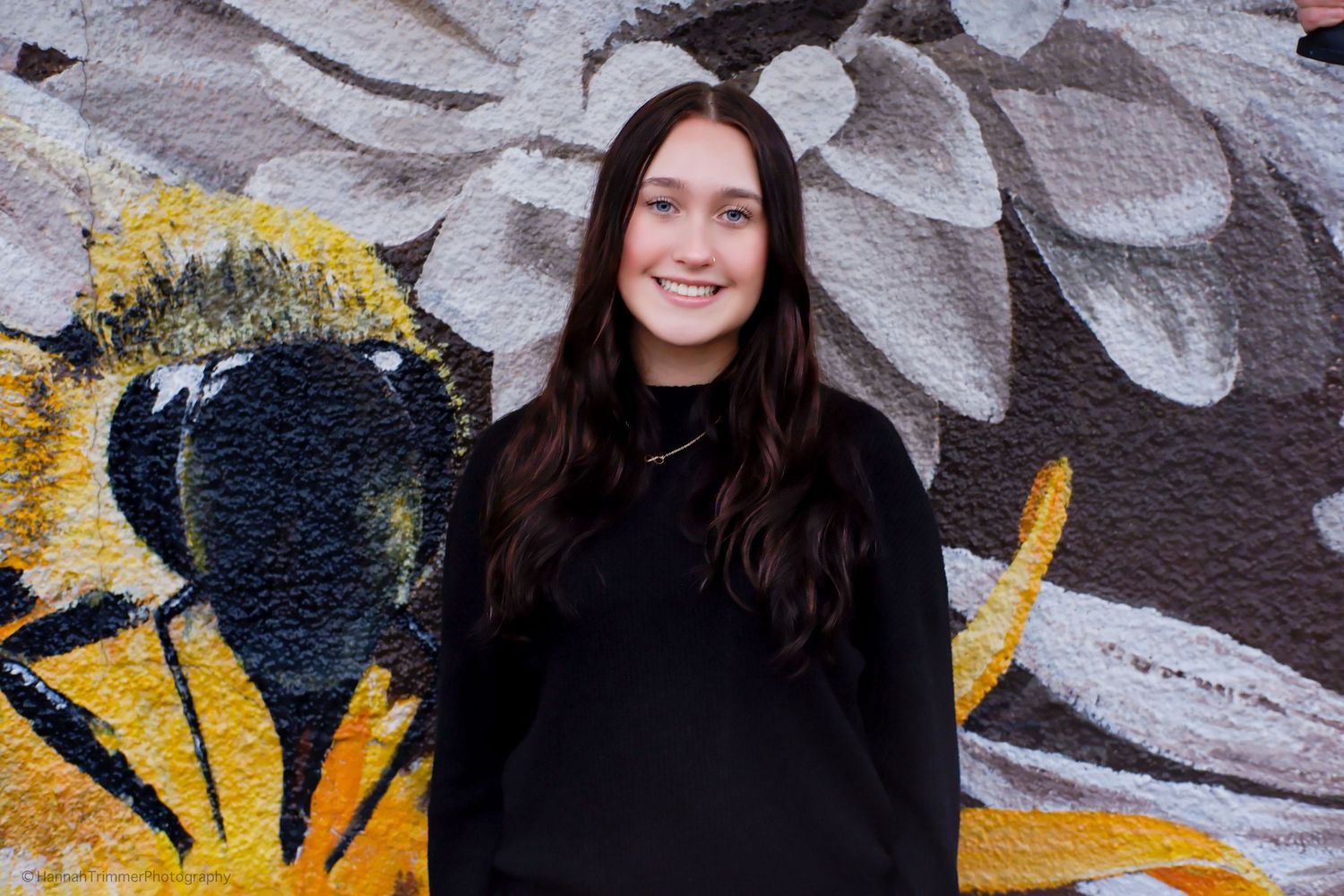 Smiling young woman in black outfit standing before colorful floral mural.