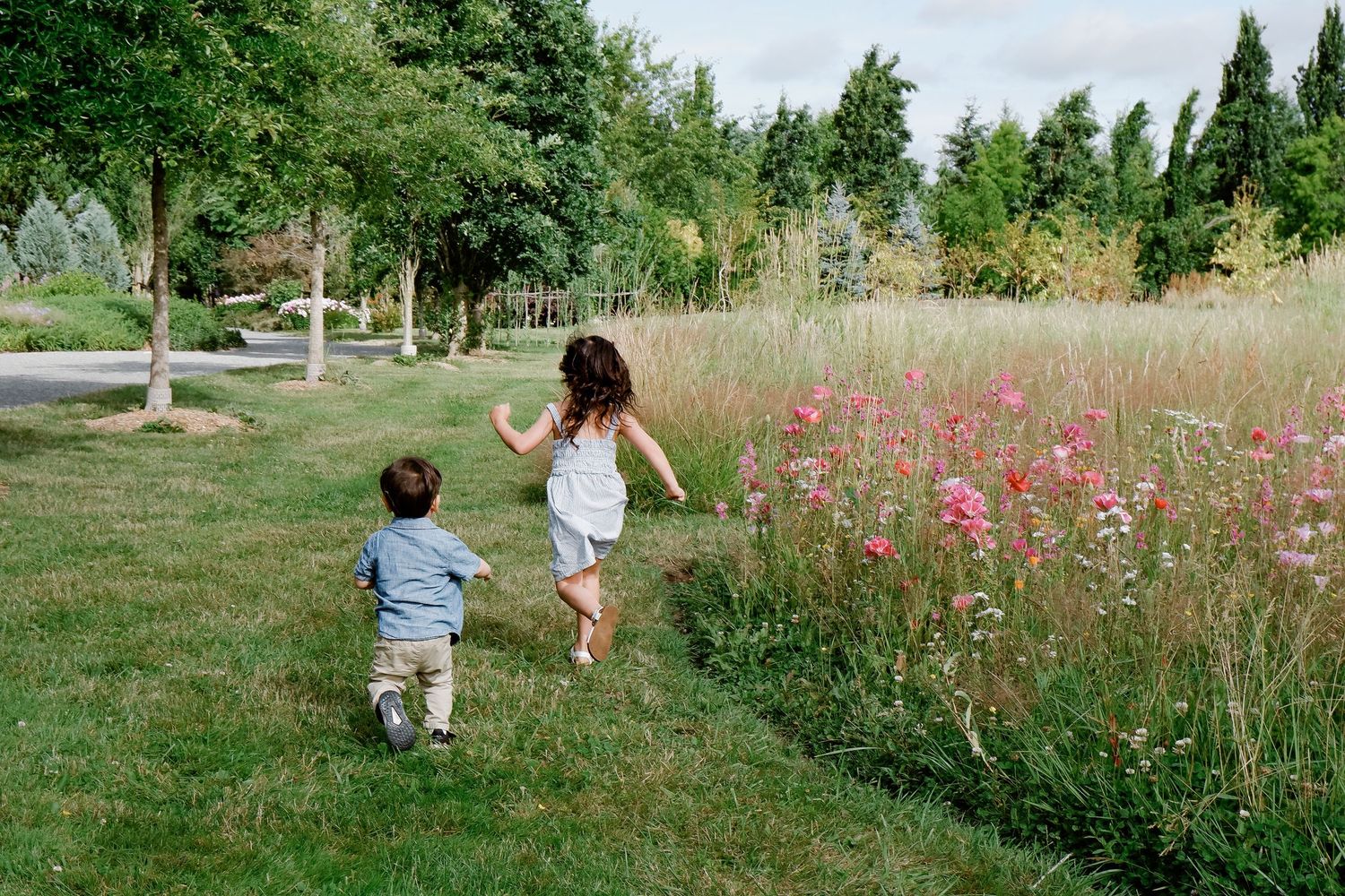 Two young children run together across a green lawn beside a field of pink wildflowers and tall trees.