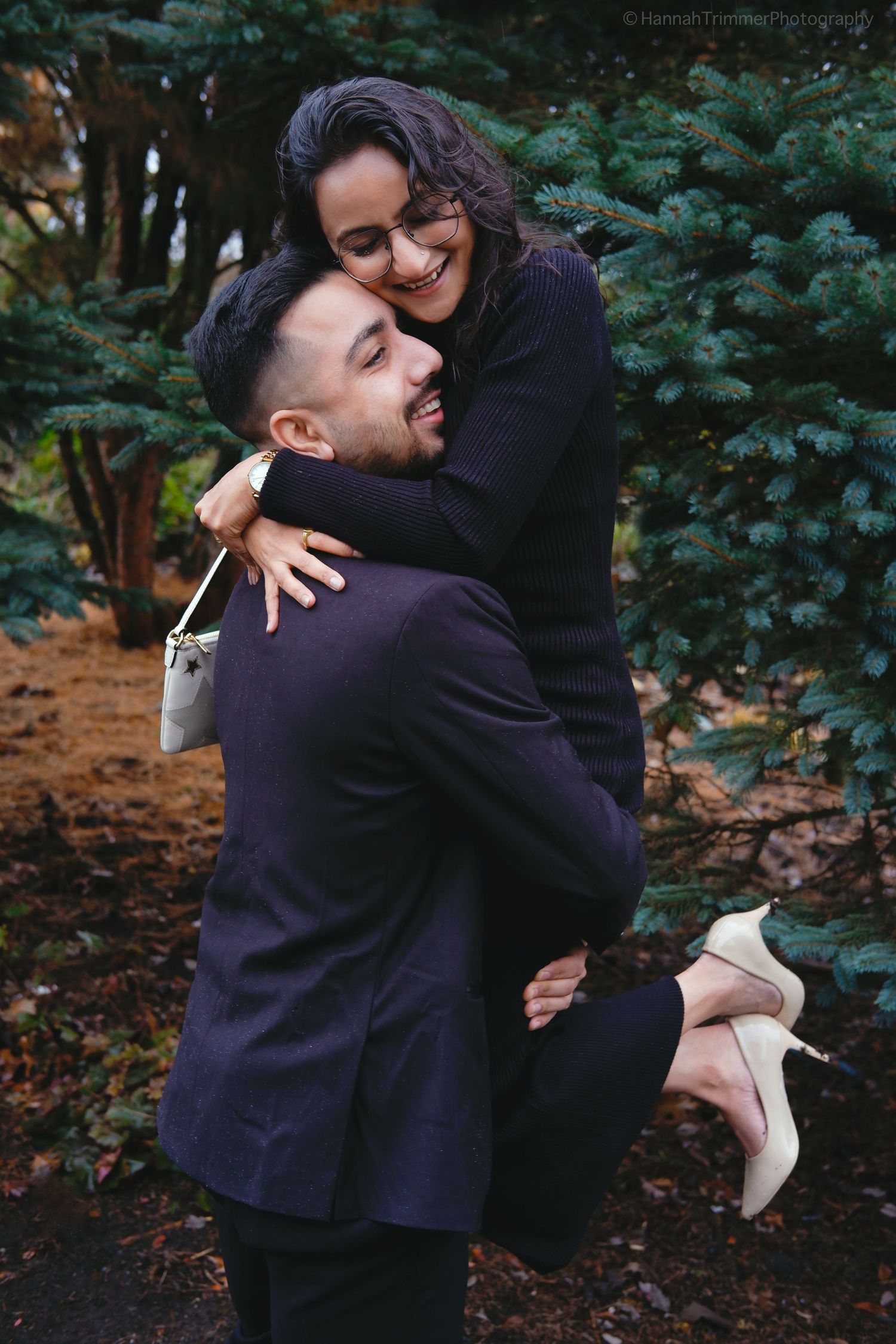 A couple embraces joyfully outdoors, man lifting woman in dark sweater dress near evergreen trees in autumn.