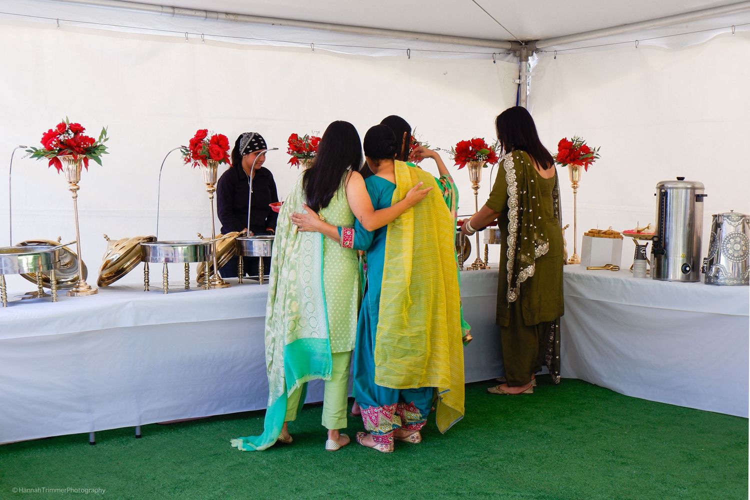 Four women in colorful salwar kameez embracing at an outdoor Indian wedding catering buffet table.