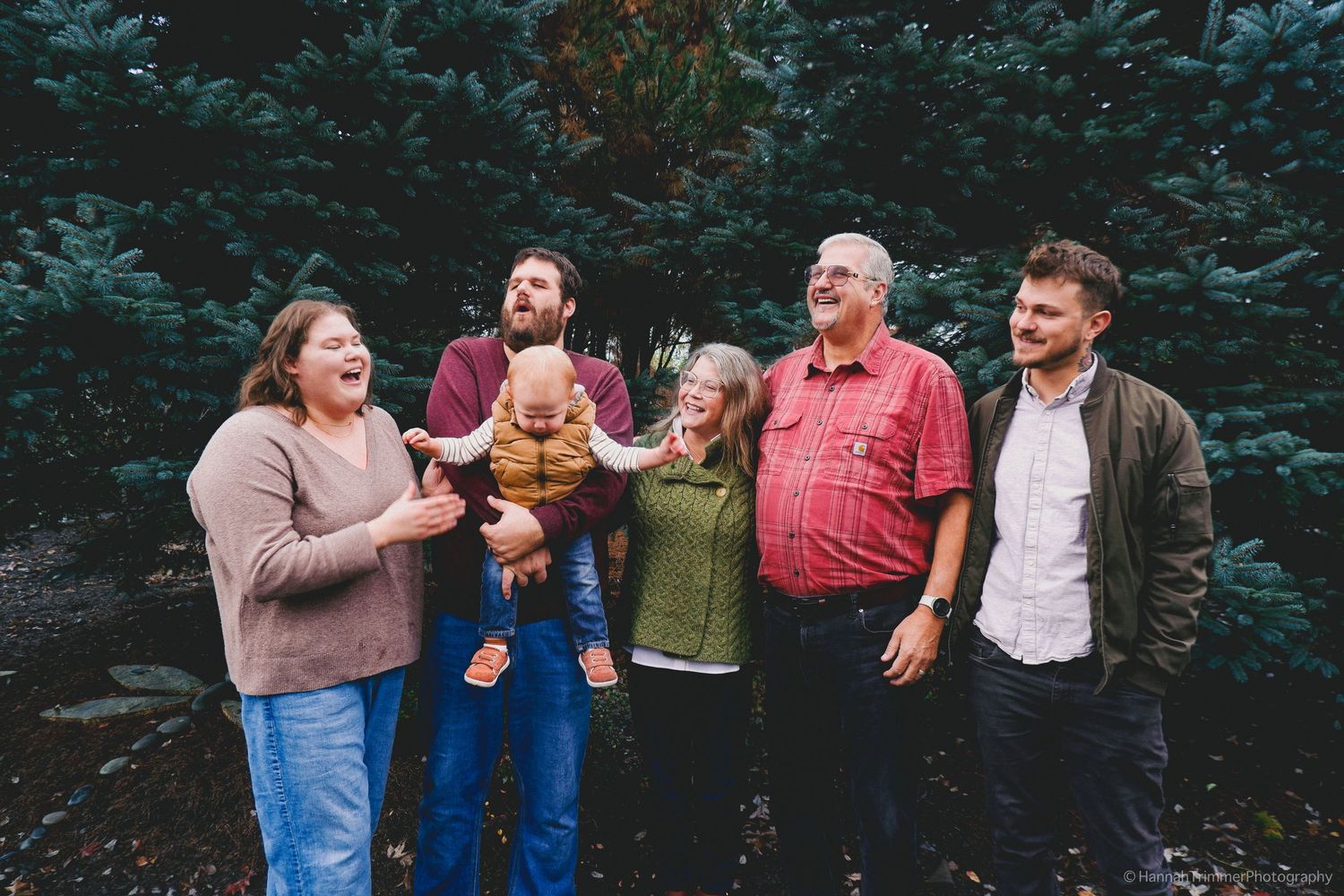 Multi-generational family of six laughing together outdoors near pine trees.