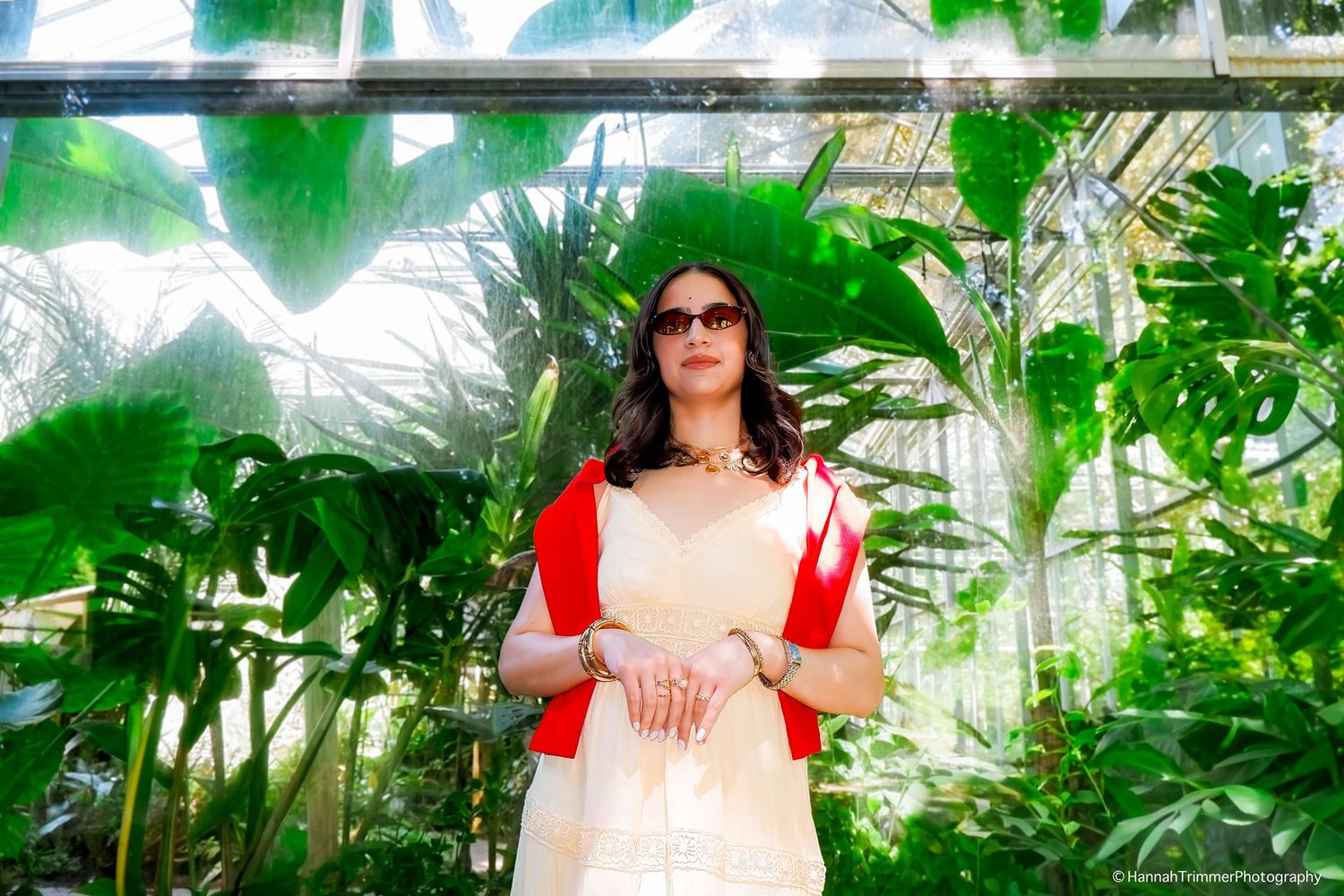 Elegant woman in cream and red outfit with sunglasses posing in tropical greenhouse garden.