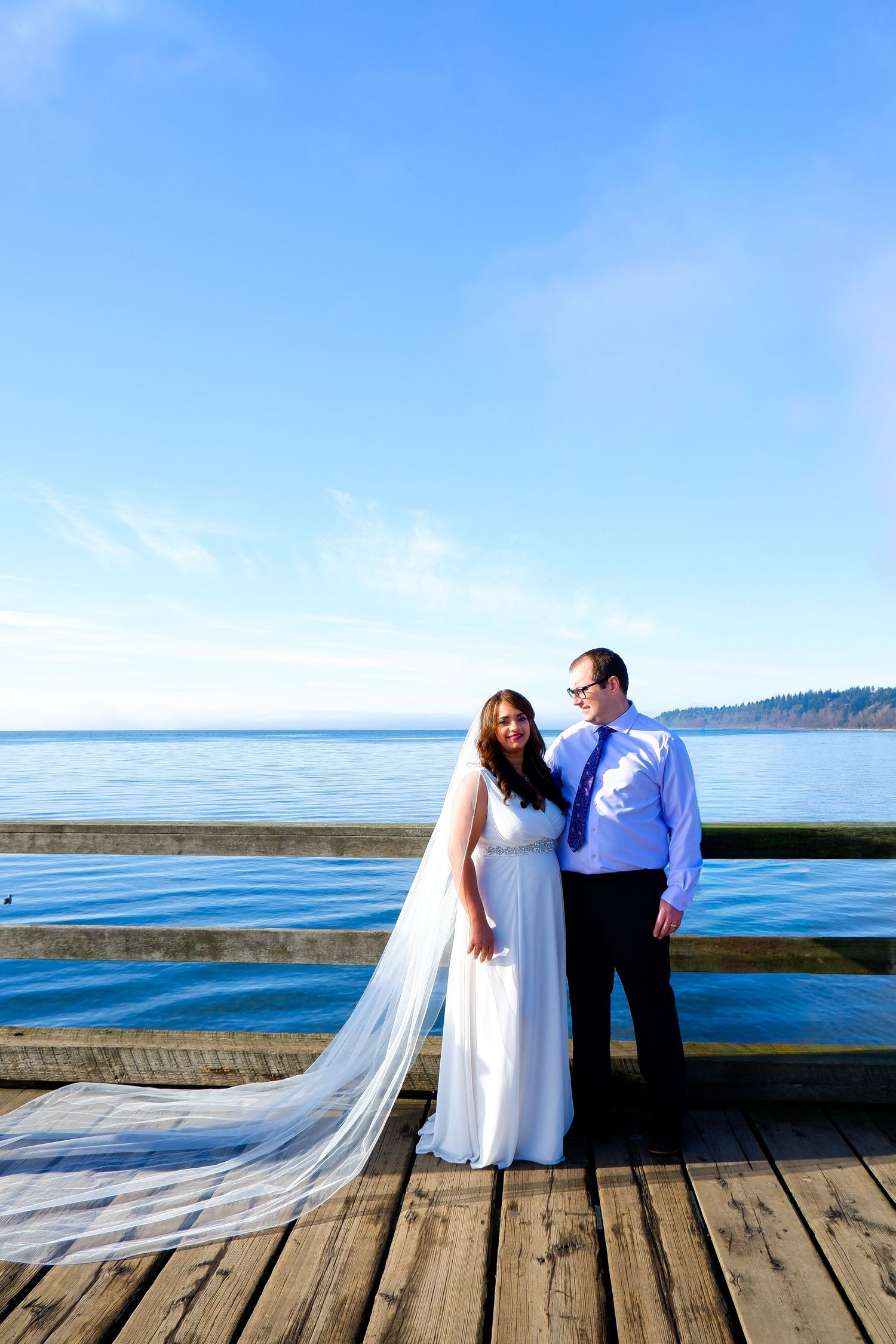 Bride in white wedding gown with long cathedral veil poses with groom on wooden pier overlooking serene blue ocean.