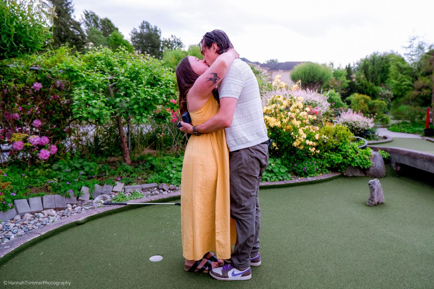 Couple sharing a romantic kiss on a mini golf course surrounded by lush colorful garden flowers.