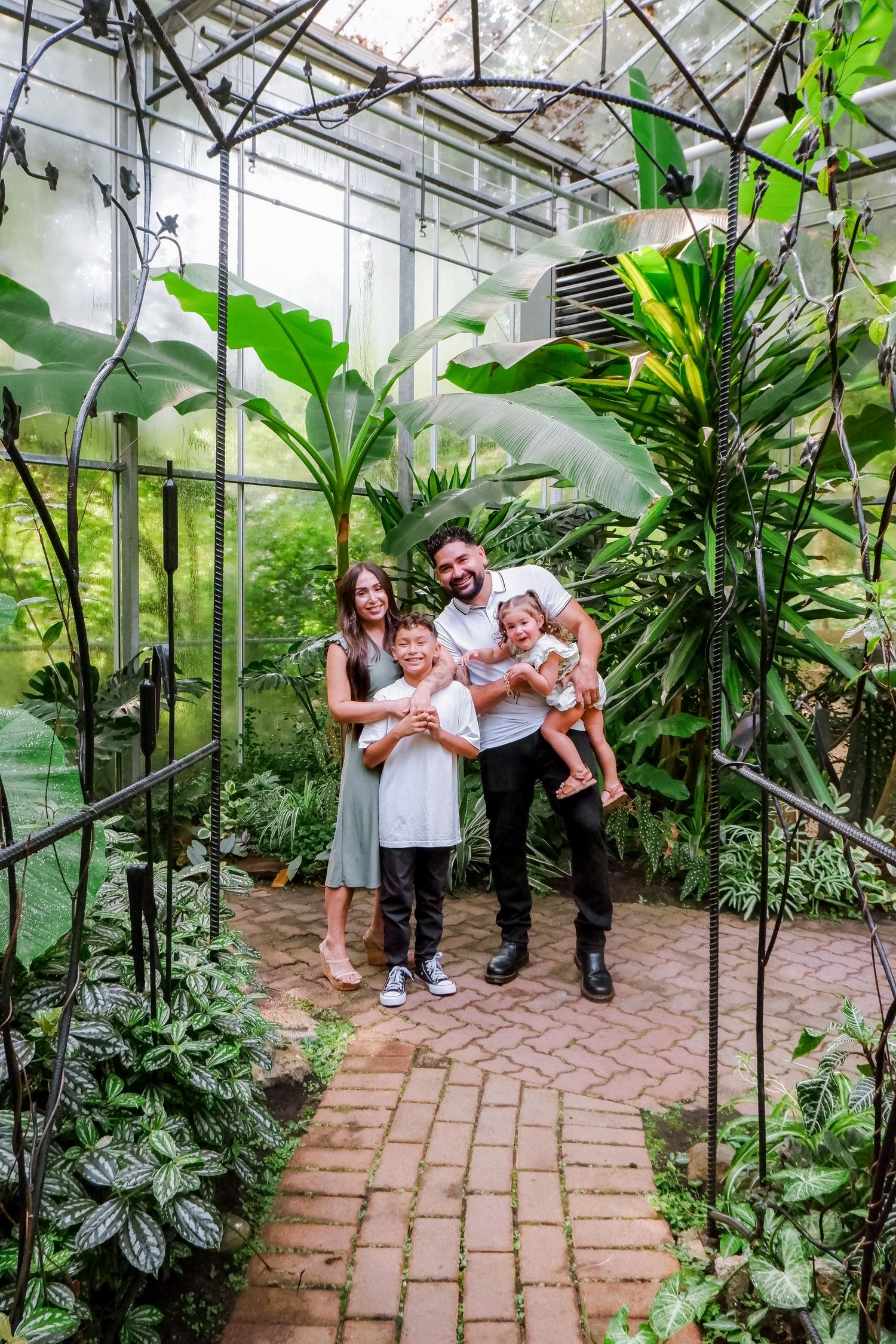 Family of four smiling together in a lush tropical greenhouse with brick path and green plants.