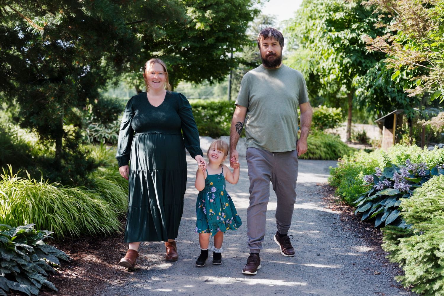 A family of three walks hand-in-hand on a garden path, parents holding their toddler girl in a green dress.