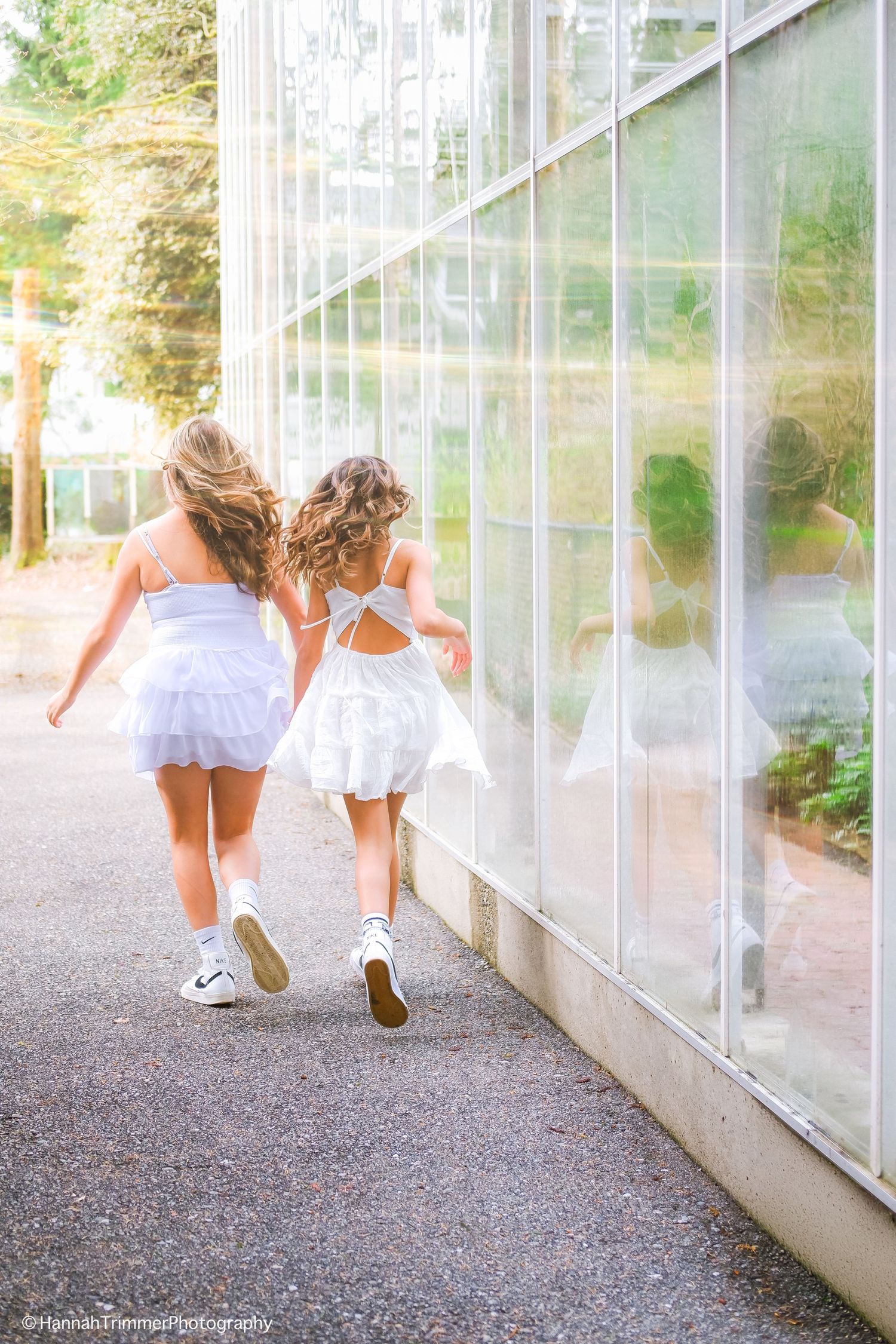 Two girls in white dresses and sneakers walk hand-in-hand along glass greenhouse wall in sunlight.