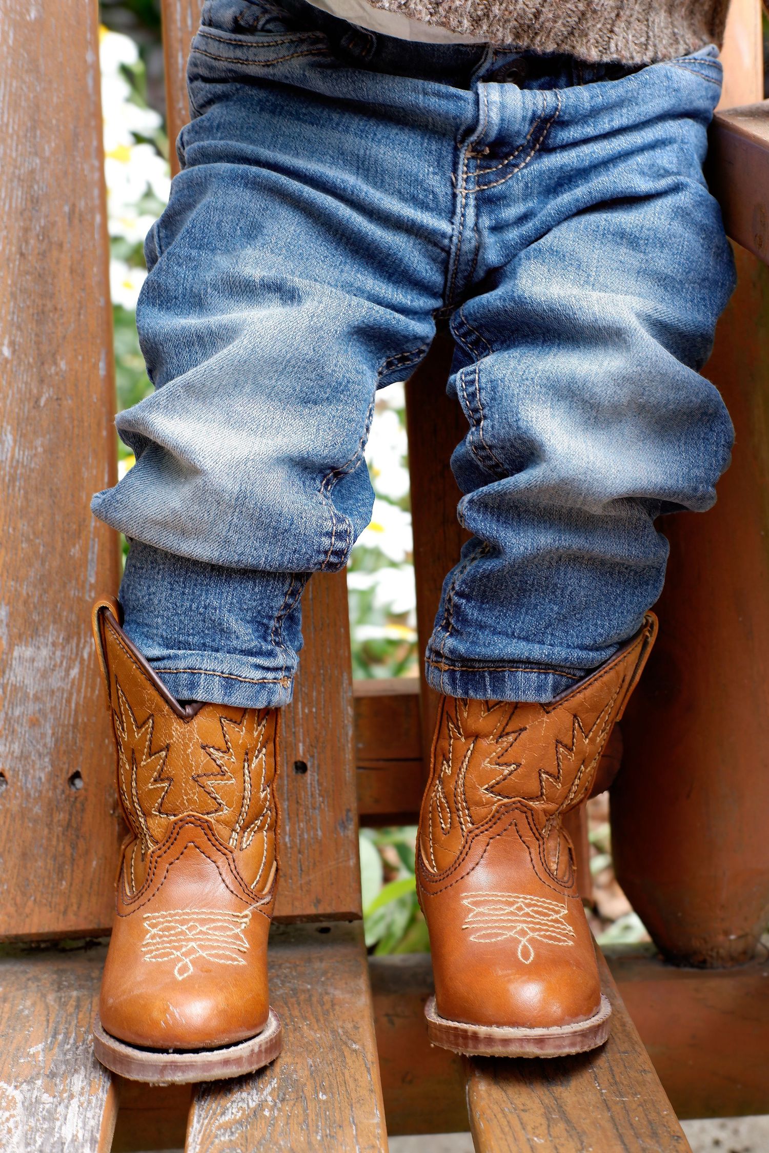 Close-up of toddler legs in jeans and brown cowboy boots standing on wooden steps outdoors.