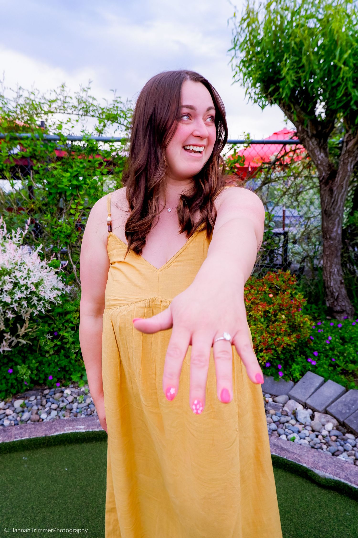 Happy woman in yellow dress showing off her engagement ring in a garden setting.