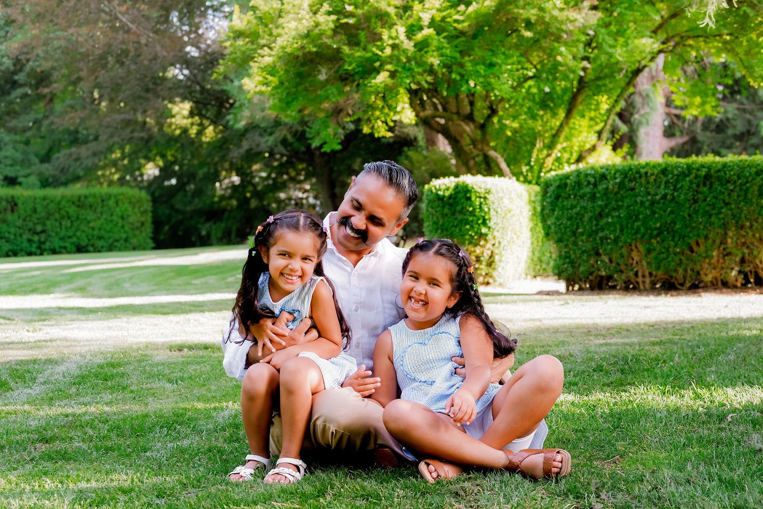 A man sits on green grass with two young girls, smiling in a sunny park with lush hedges and trees.