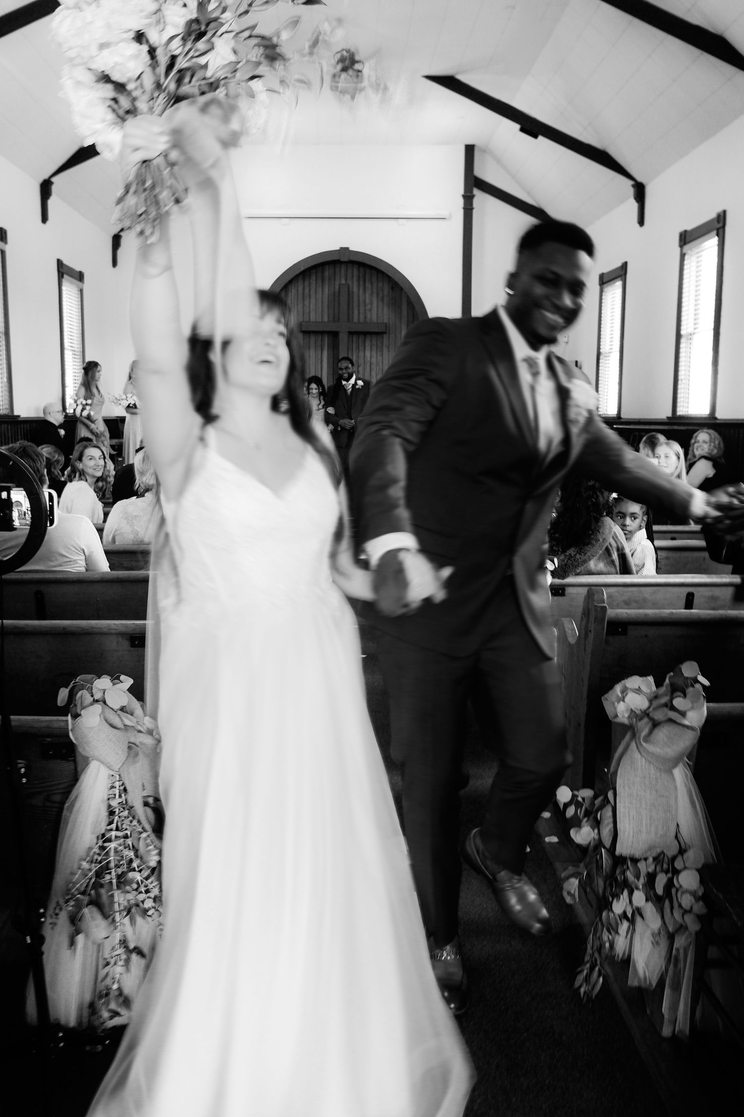 Black and white photo of joyful newlyweds celebrating recessional in chapel, bride raising bouquet.