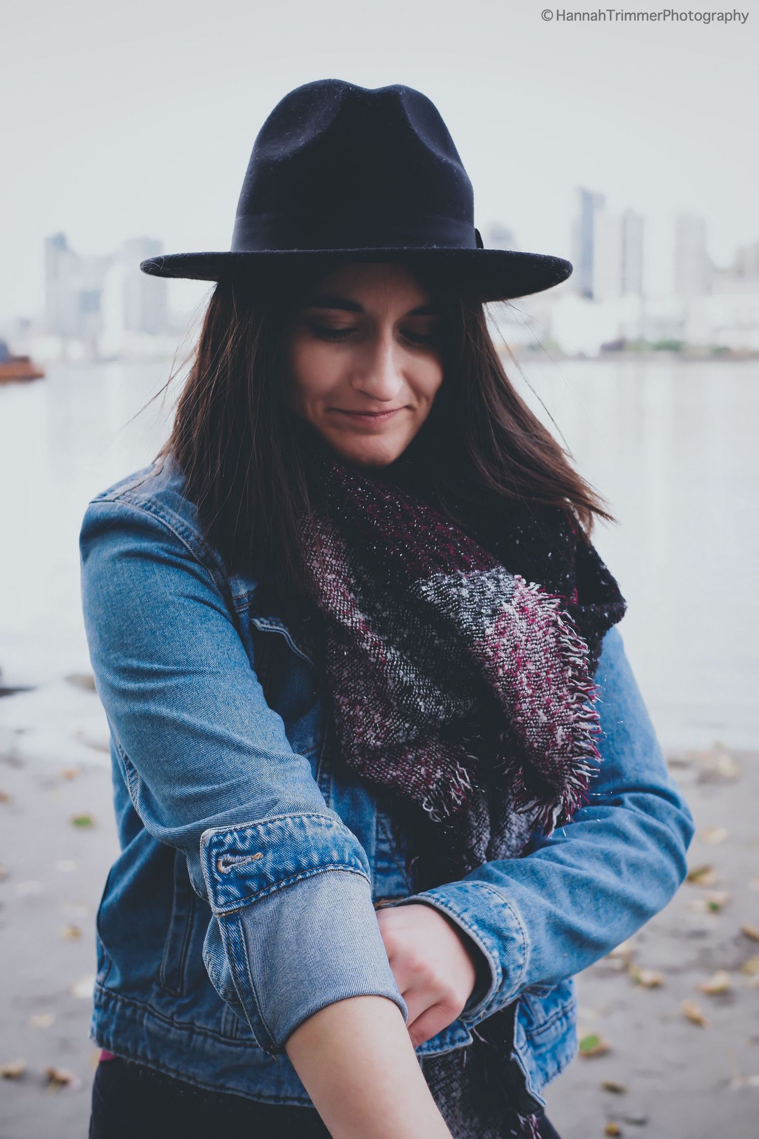Woman in black fedora and denim jacket looking down near waterfront on overcast day.