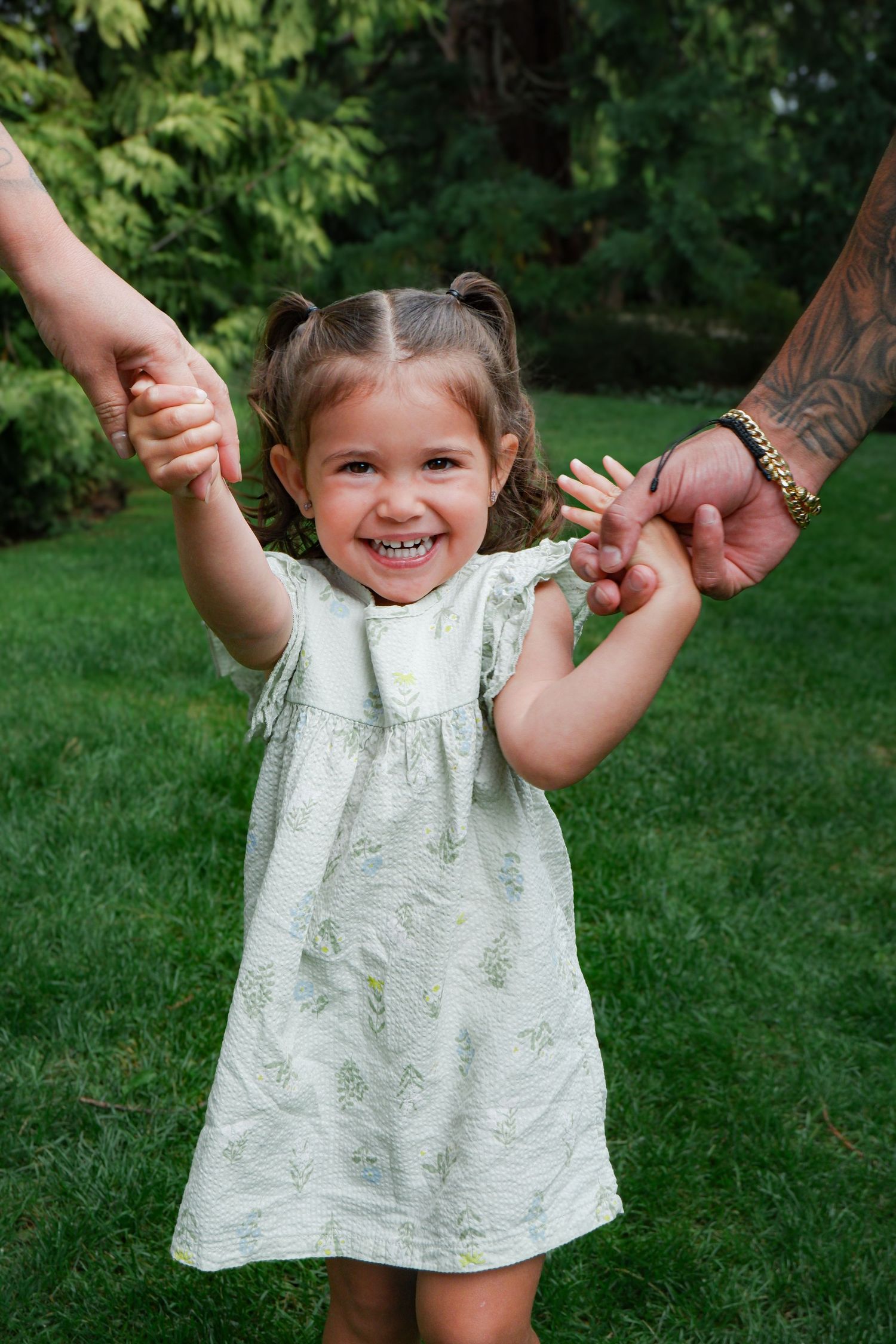 A joyful toddler girl in a green floral dress holds hands with two adults outdoors on a lush green lawn.