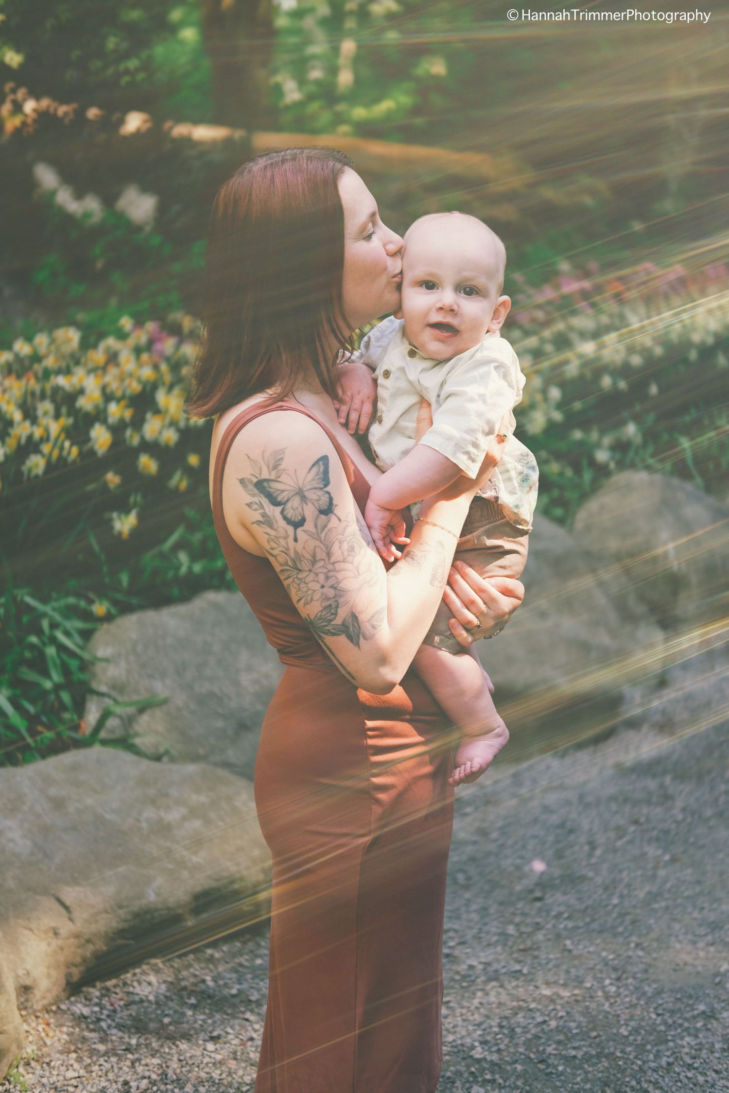 Tattooed mother kissing her baby in a sunlit garden setting with soft bokeh background.