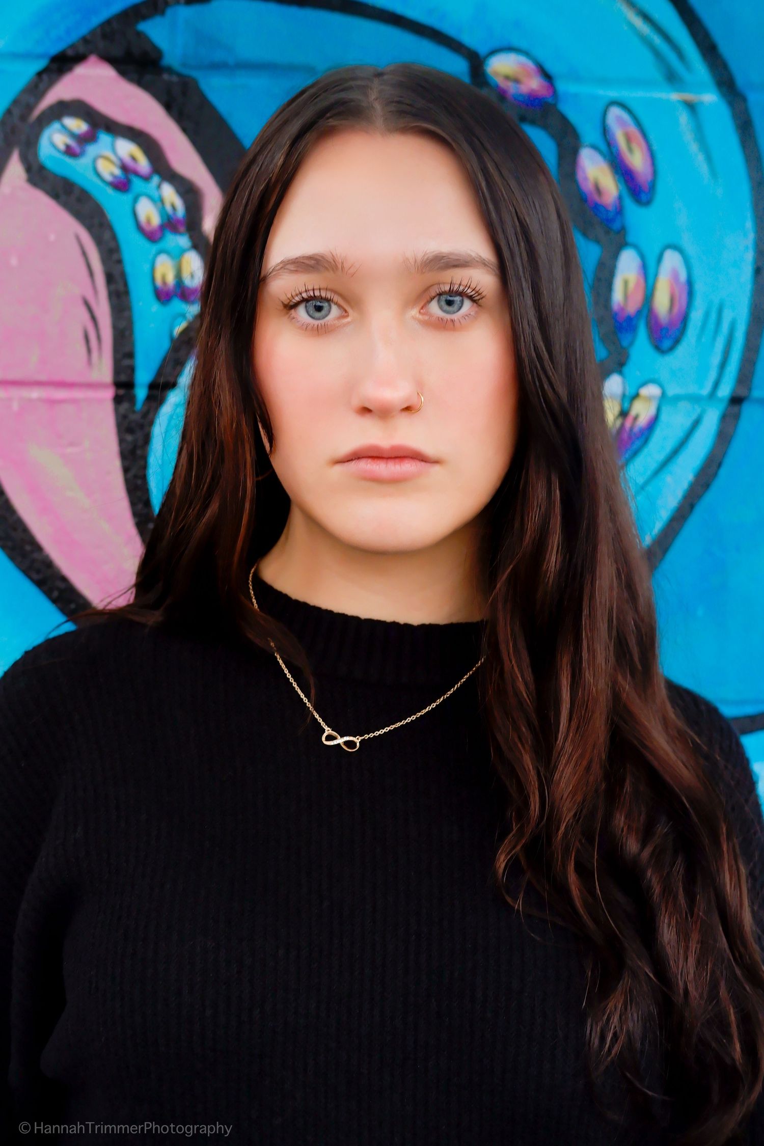 Teen girl with blue eyes in black sweater posing before colorful graffiti wall.