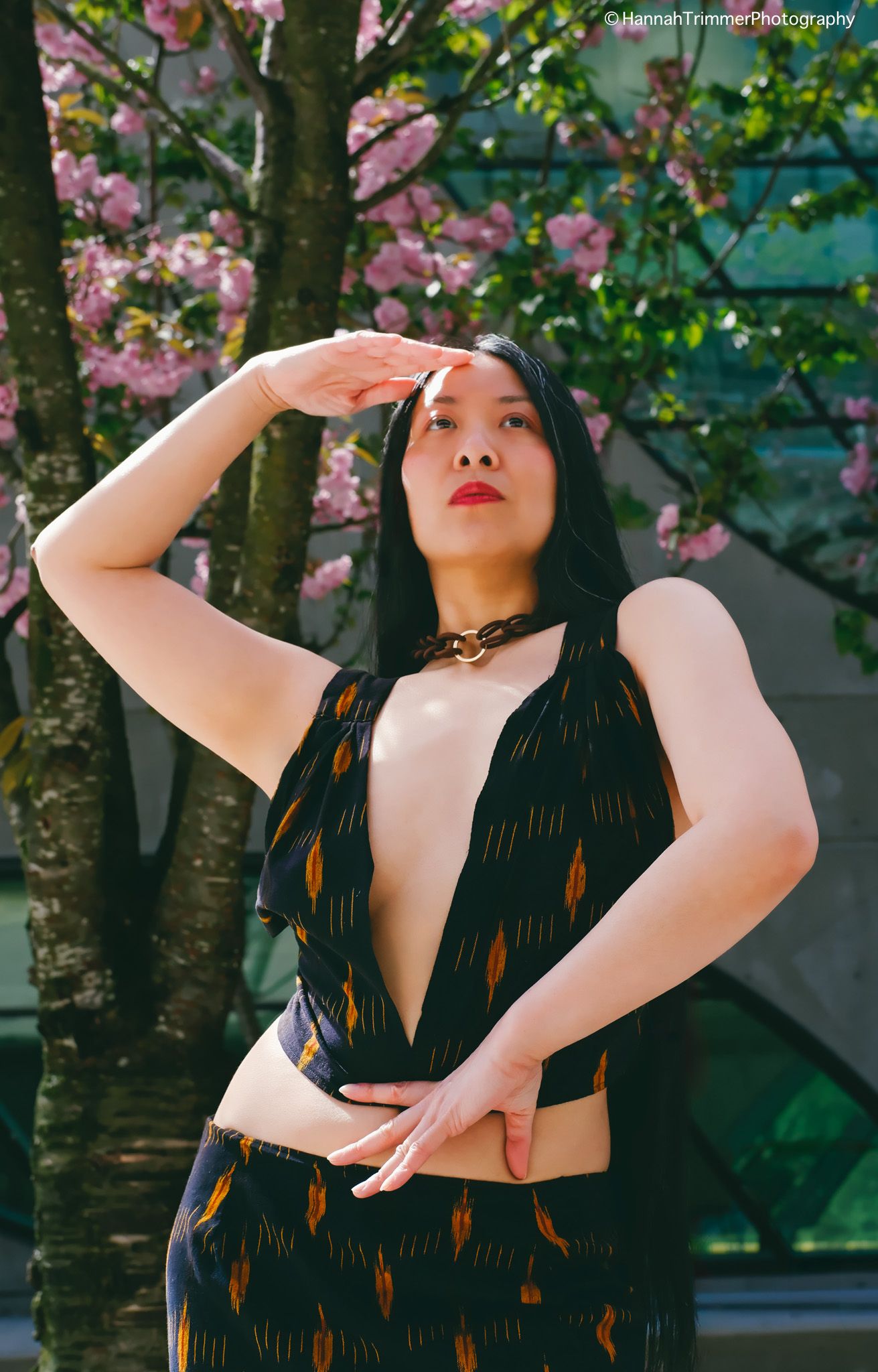 Woman in a deep V-neck patterned dress posing under pink cherry blossom trees in an outdoor setting.