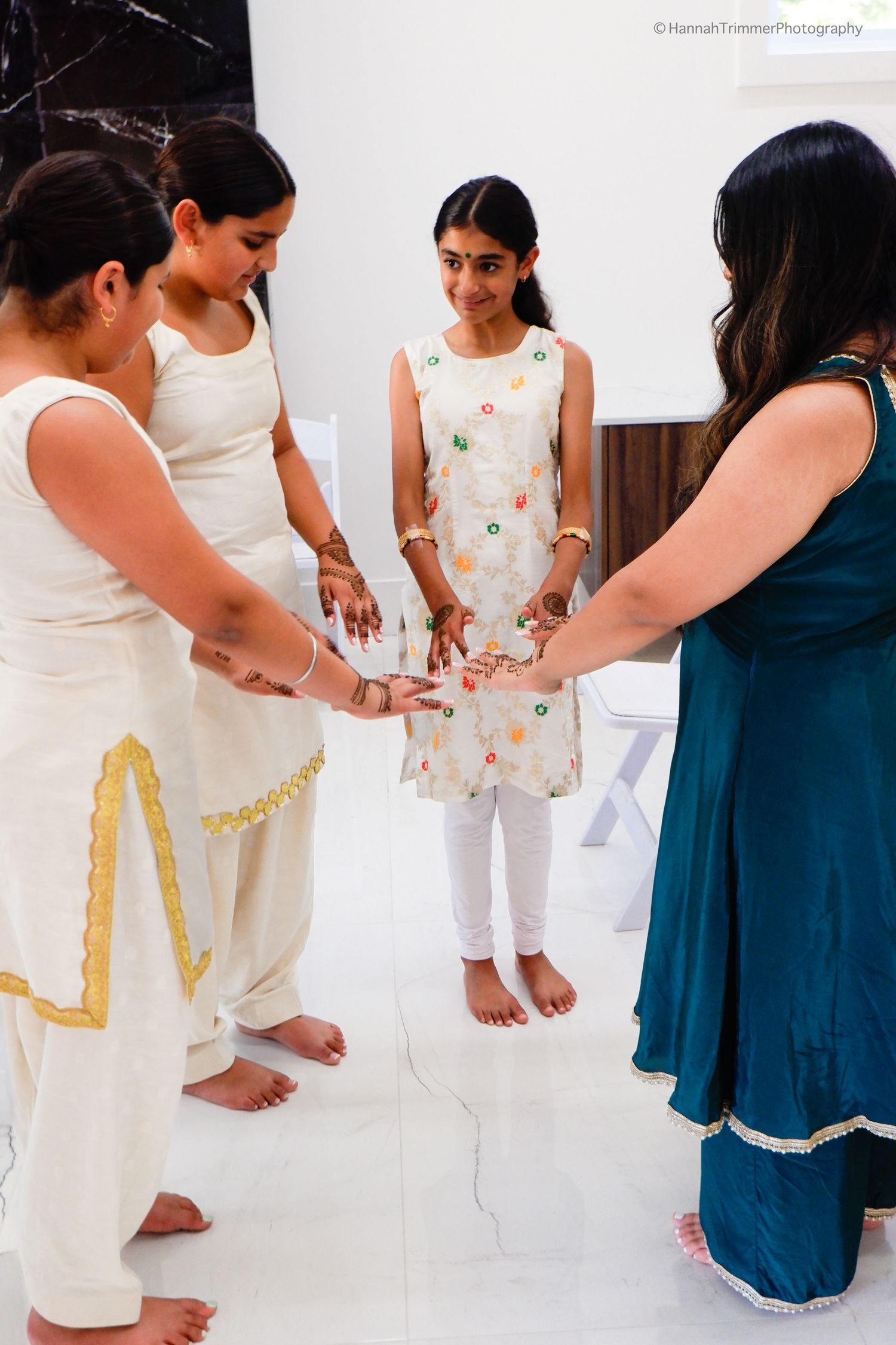 Young girls in white traditional Indian attire join hands in a circle during a cultural ceremony indoors.