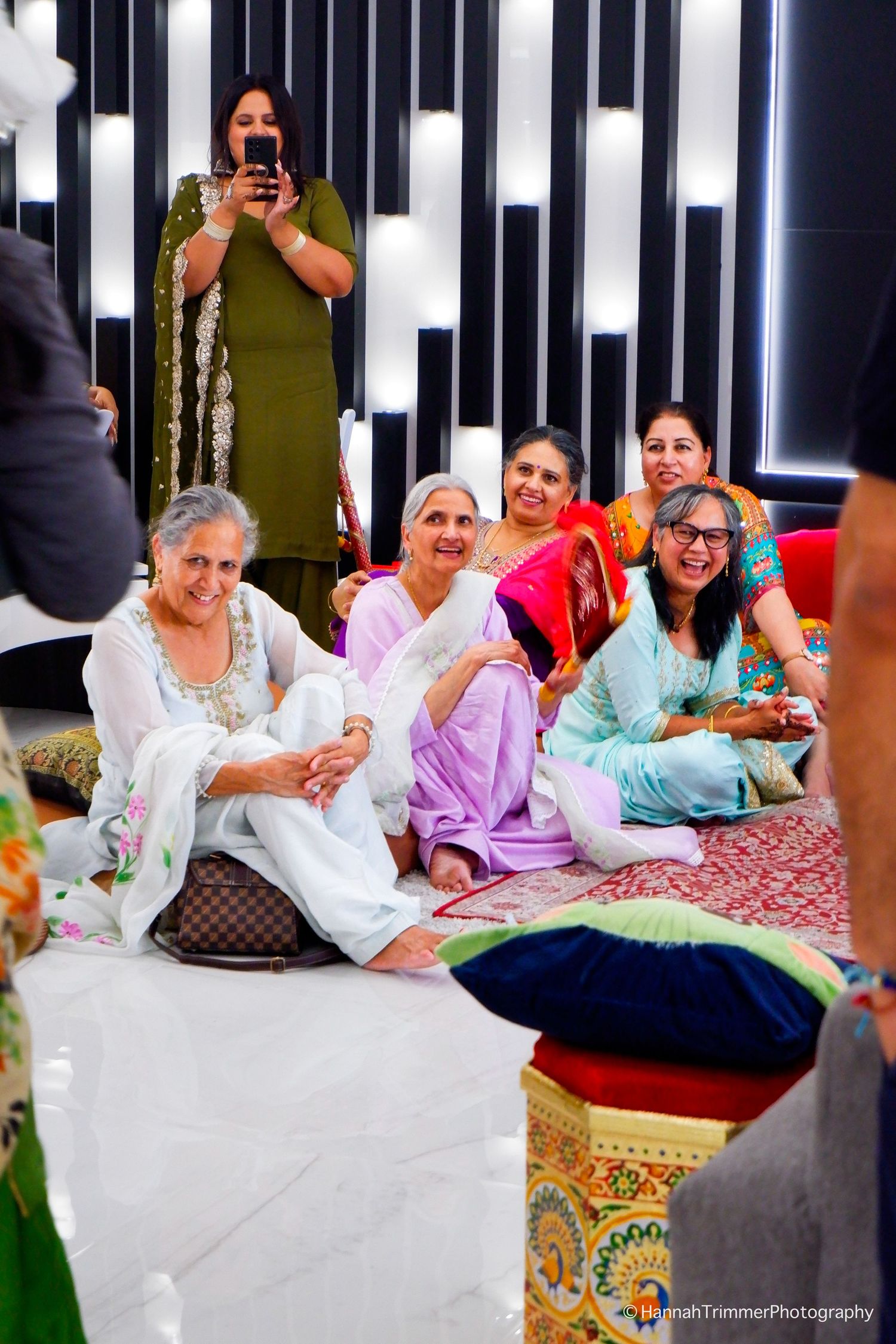 Smiling elderly women in white and pastel salwar kameez seated together at an Indian wedding celebration.
